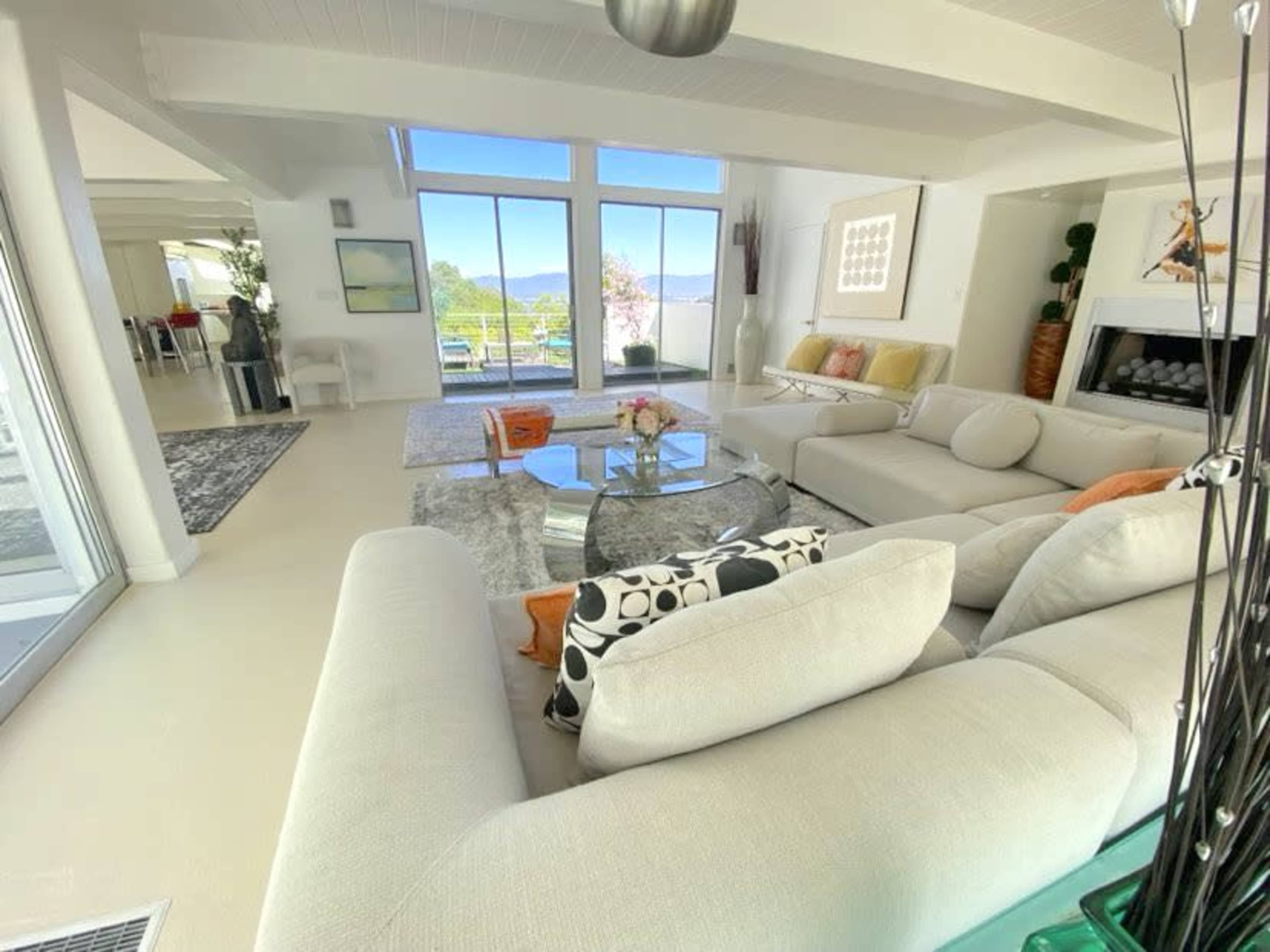 A bright, modern living room features a large sofa, a glass coffee table, and panoramic views of mountains through floor-to-ceiling windows.