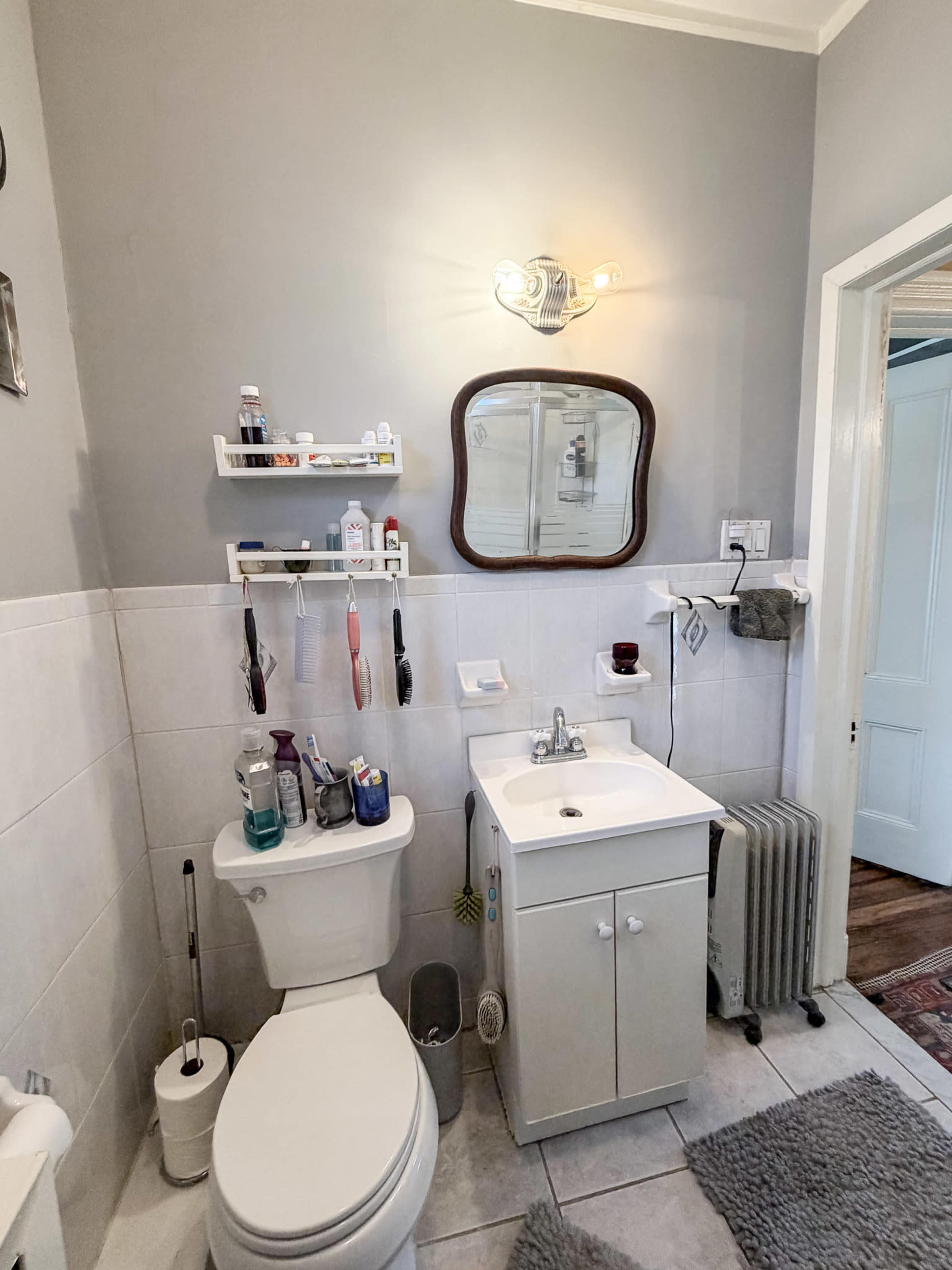 The image shows a small bathroom featuring a toilet, sink with a cabinet, and a mirror mounted on the wall, along with several toiletries on shelves.