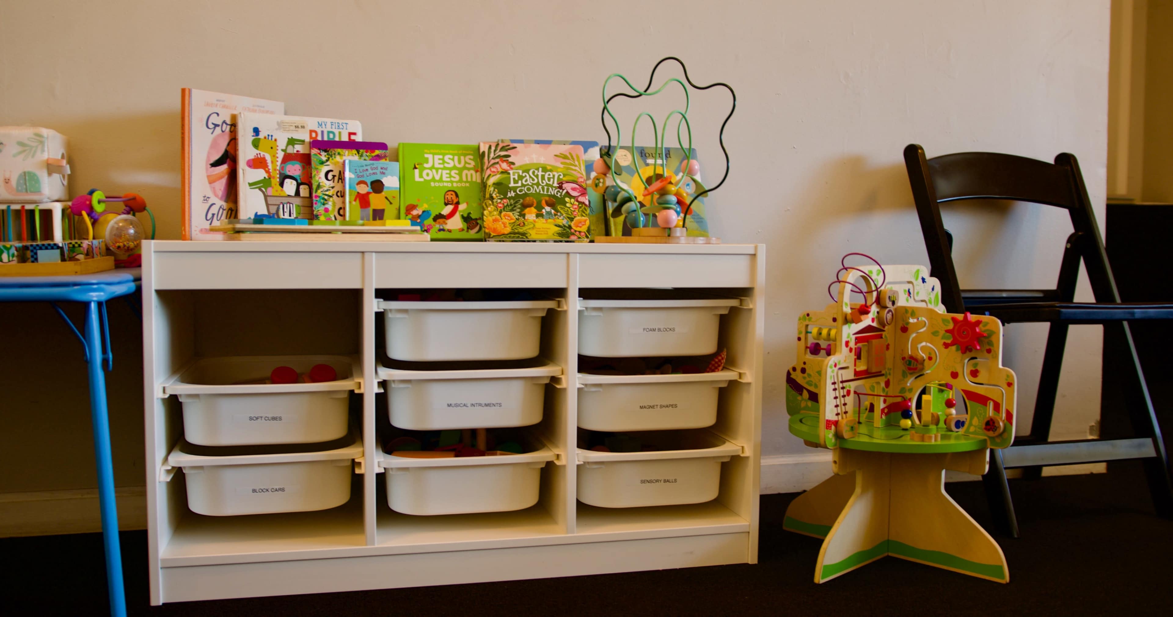 The image shows a storage unit with labeled bins filled with toys, alongside a collection of children's books on top, and a colorful activity center on the side.