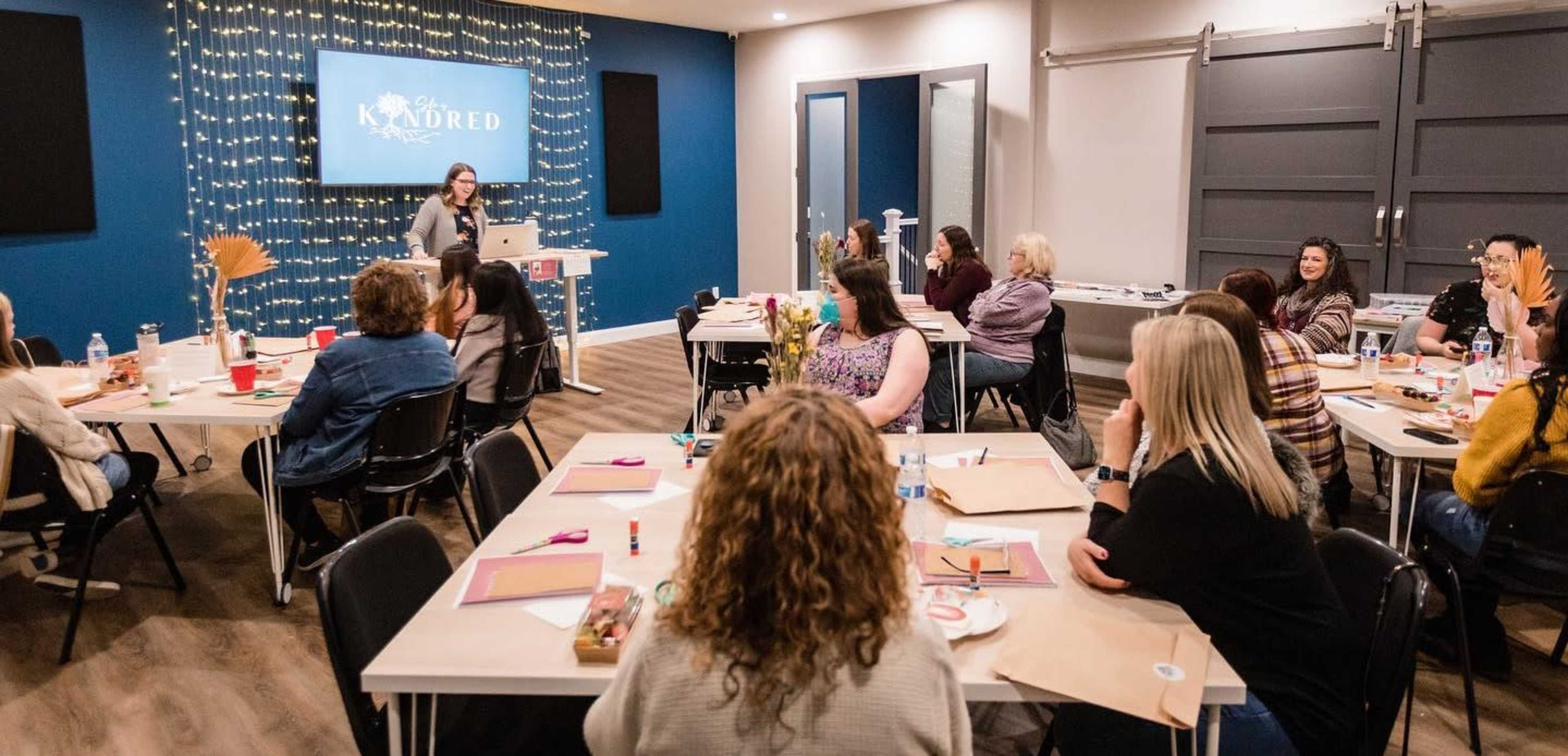 A group of people sits around tables in a well-lit room, watching a speaker present in front of a screen adorned with a decorative backdrop.
