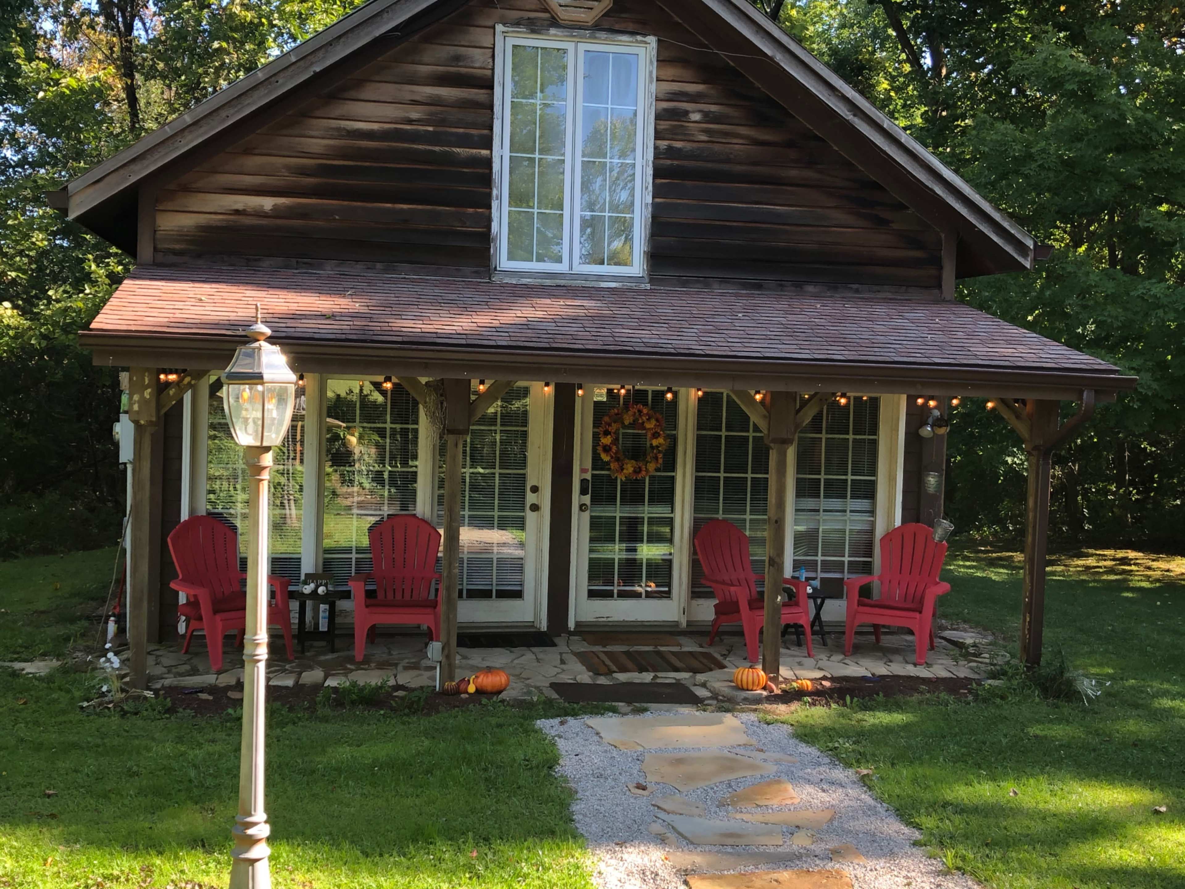 A wooden house with a front porch features red Adirondack chairs and decorative pumpkins, surrounded by grass and trees.