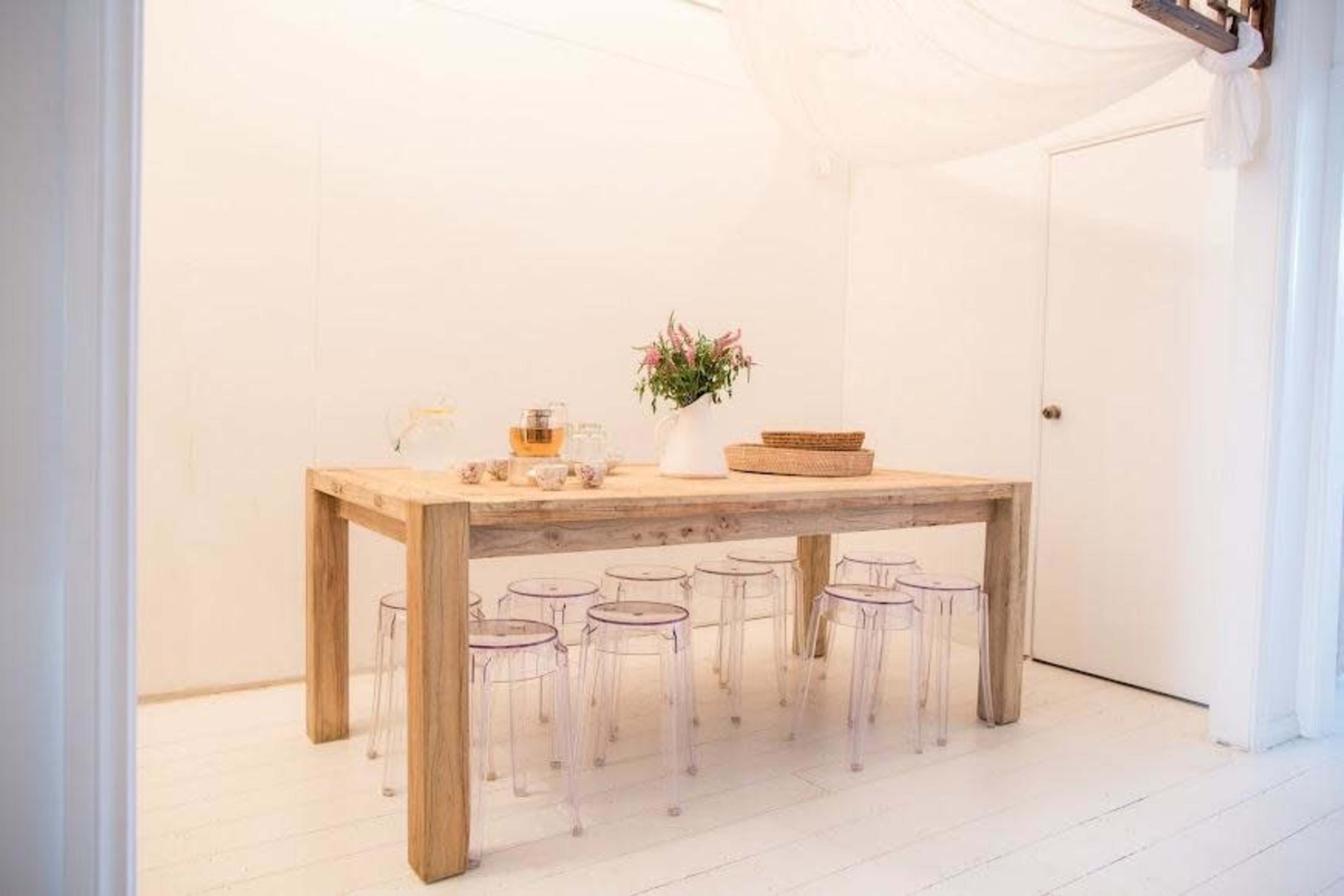 A wooden table with clear acrylic chairs is set in a bright, minimalist dining room featuring a vase of flowers.