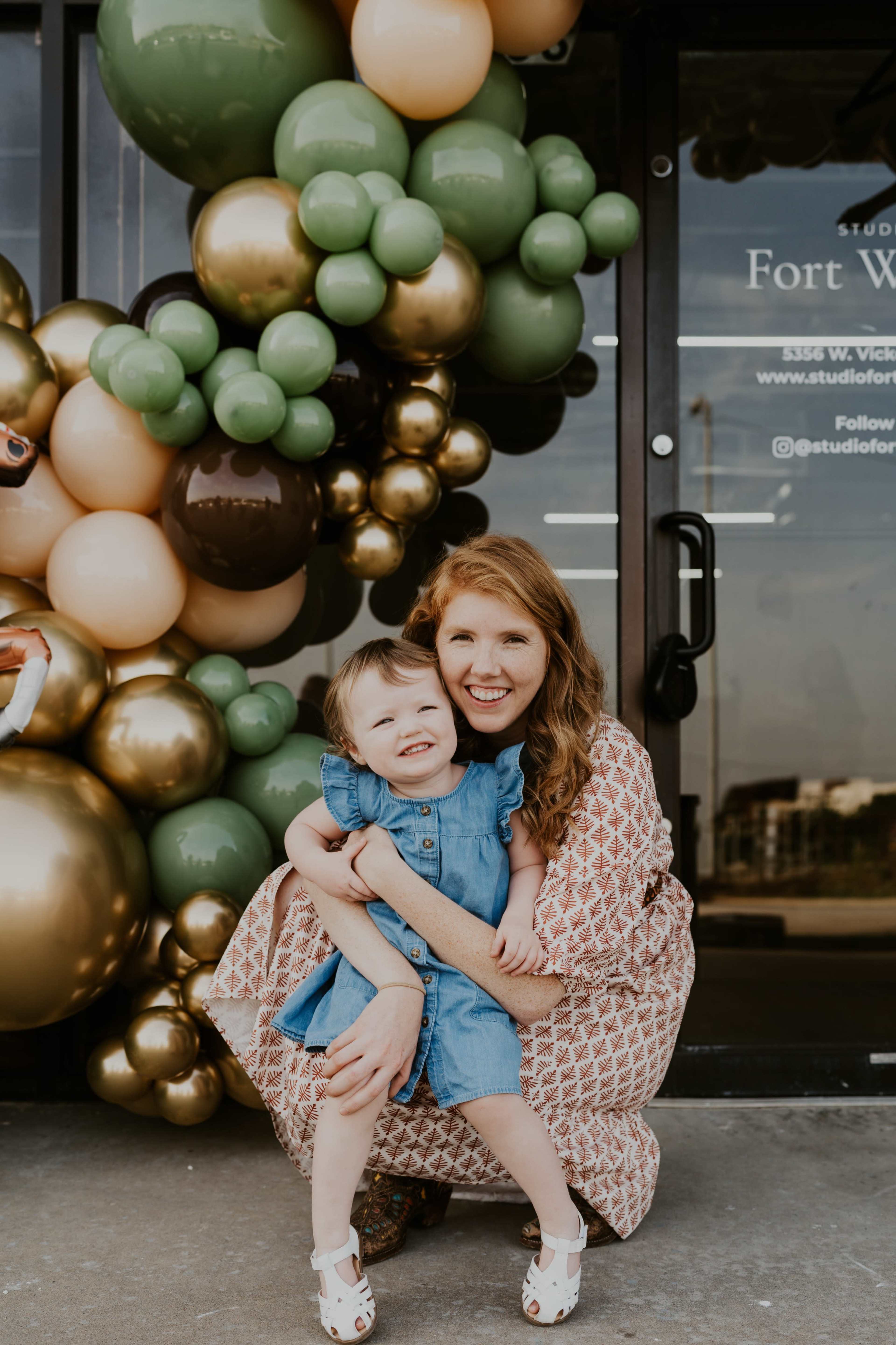 A woman in a patterned dress sits on the ground with a young girl in a blue dress, both smiling in front of a balloon arch at a storefront.