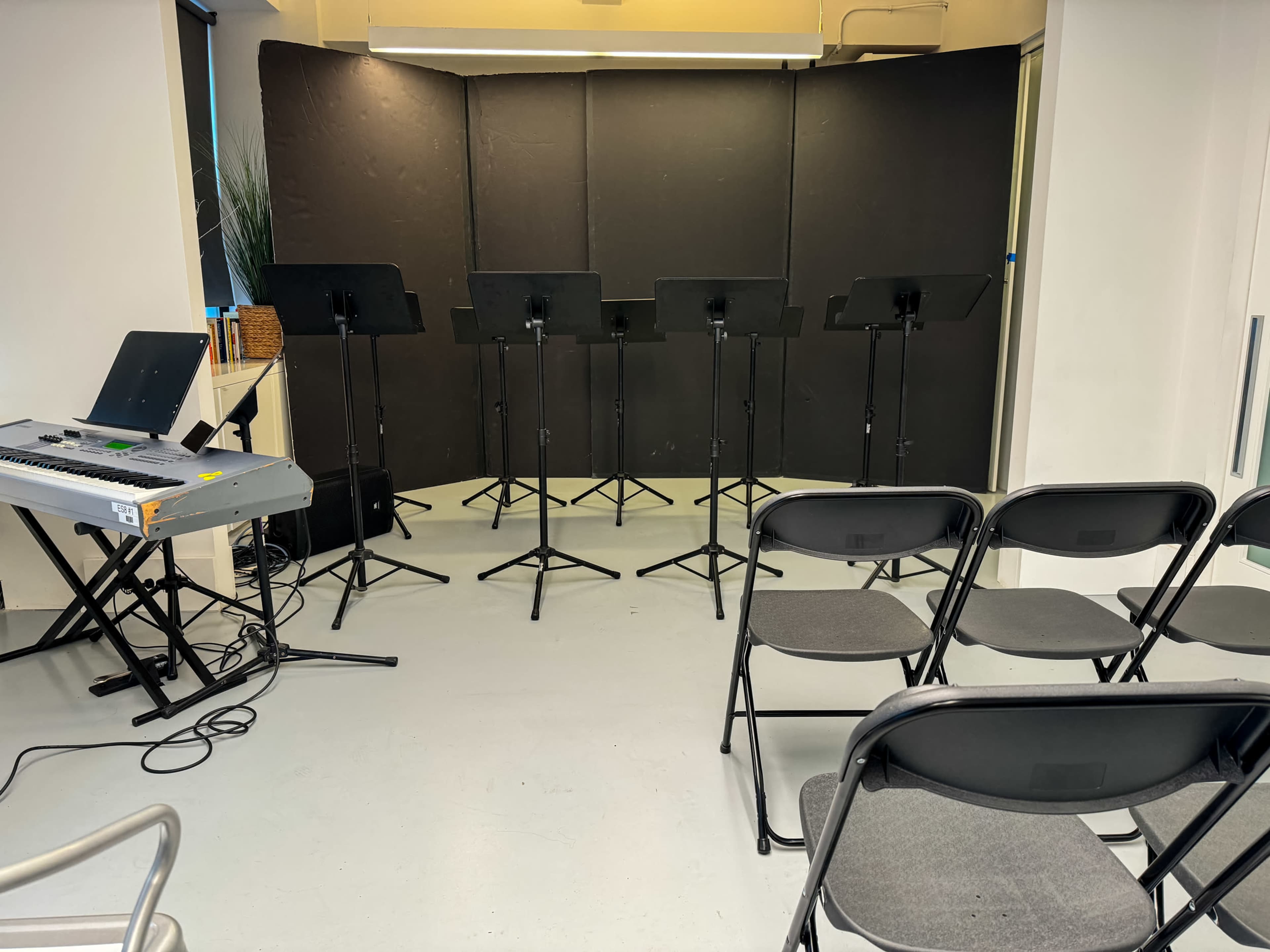 A row of music stands is set up in front of empty chairs in a rehearsal space.