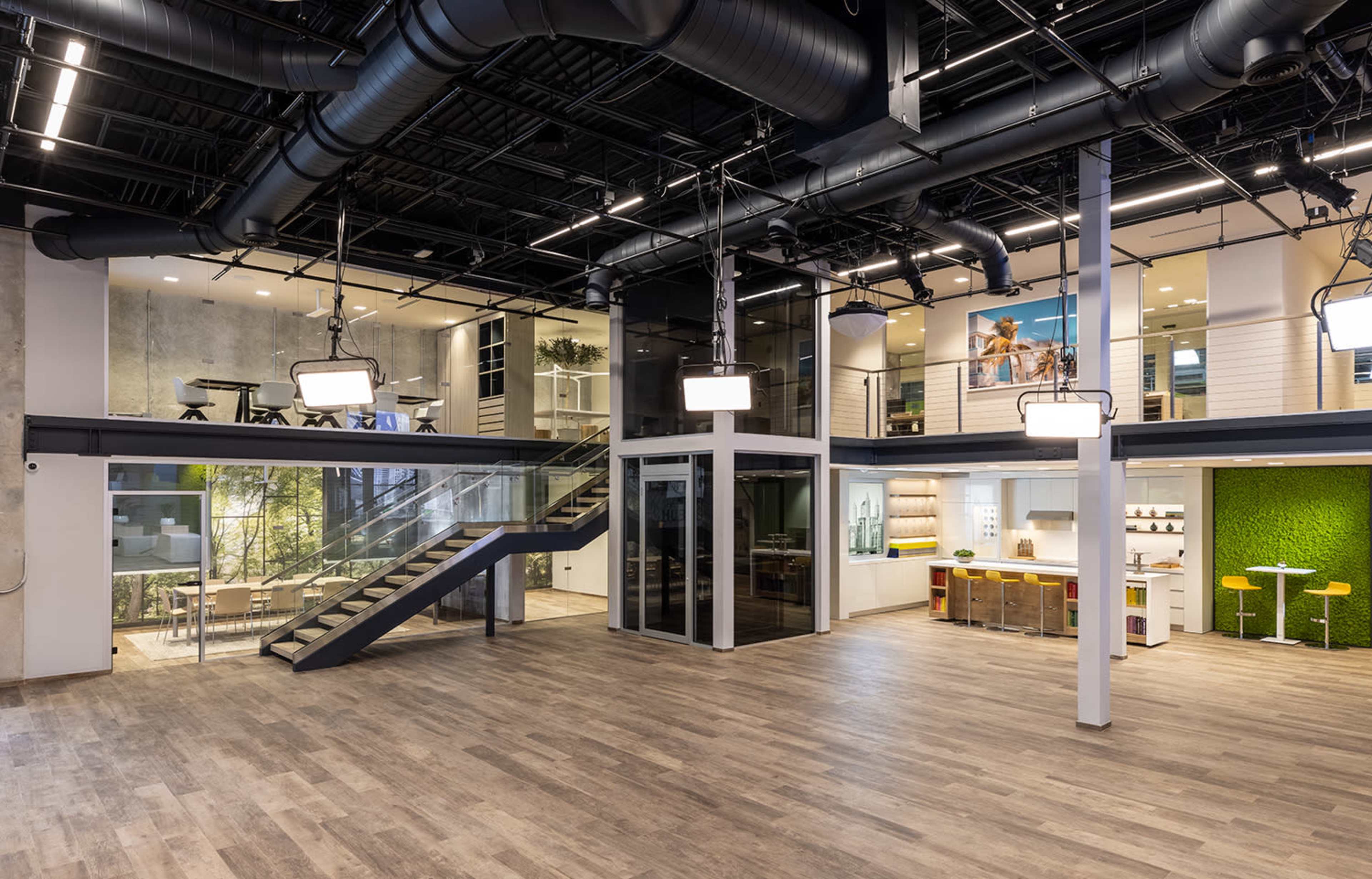 The image shows a modern office space with an open layout, featuring a staircase, glass-walled meeting rooms, and areas for collaboration and relaxation.