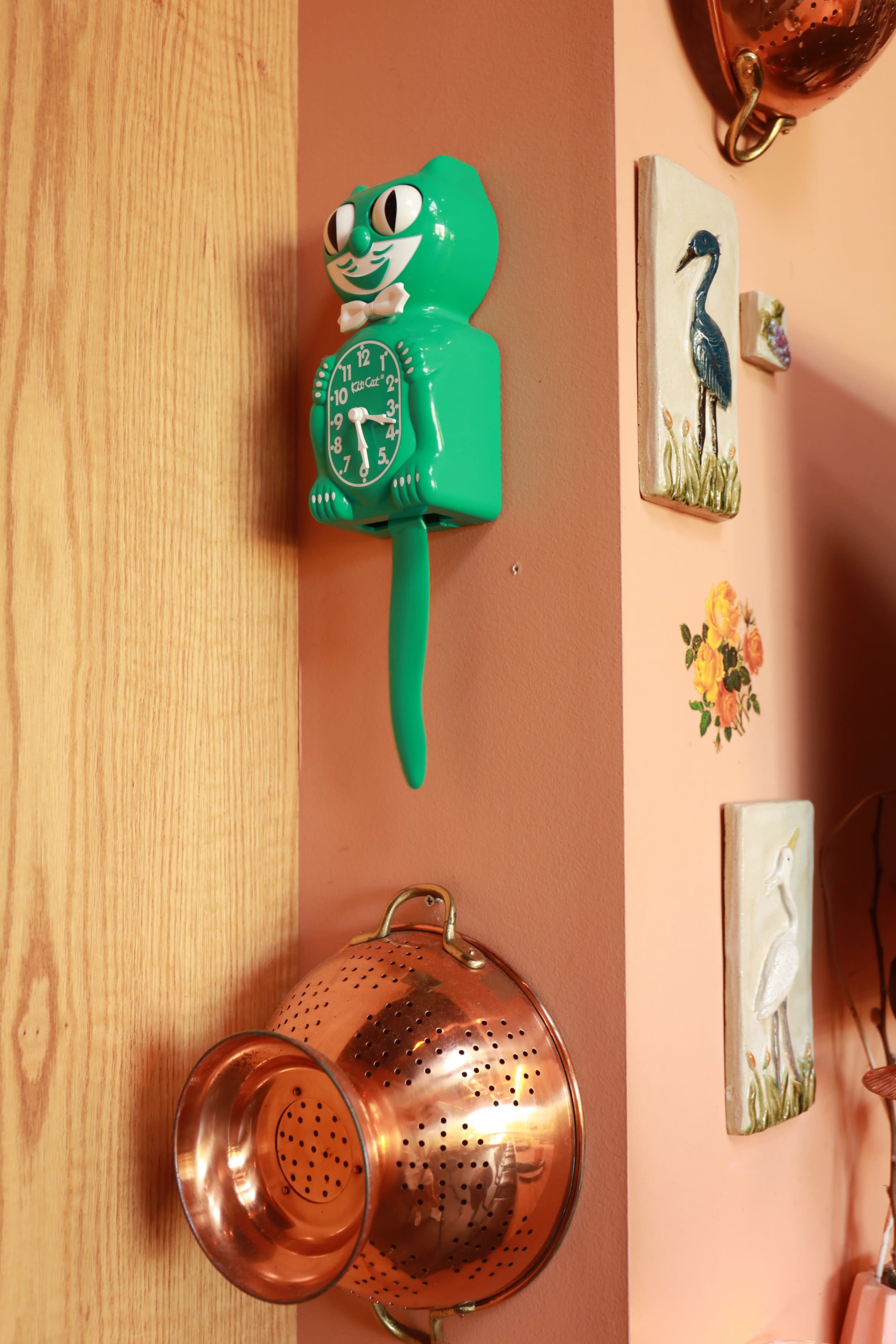 A green cat-shaped wall clock hangs near a copper colander on a decorated wall.