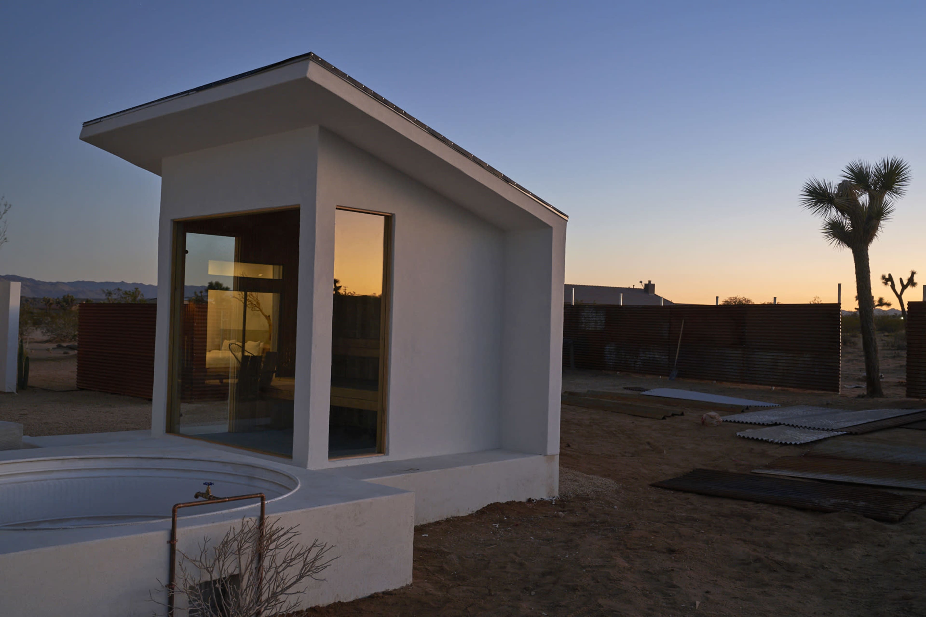 A modern white structure with large windows is situated in a desert landscape at sunset.
