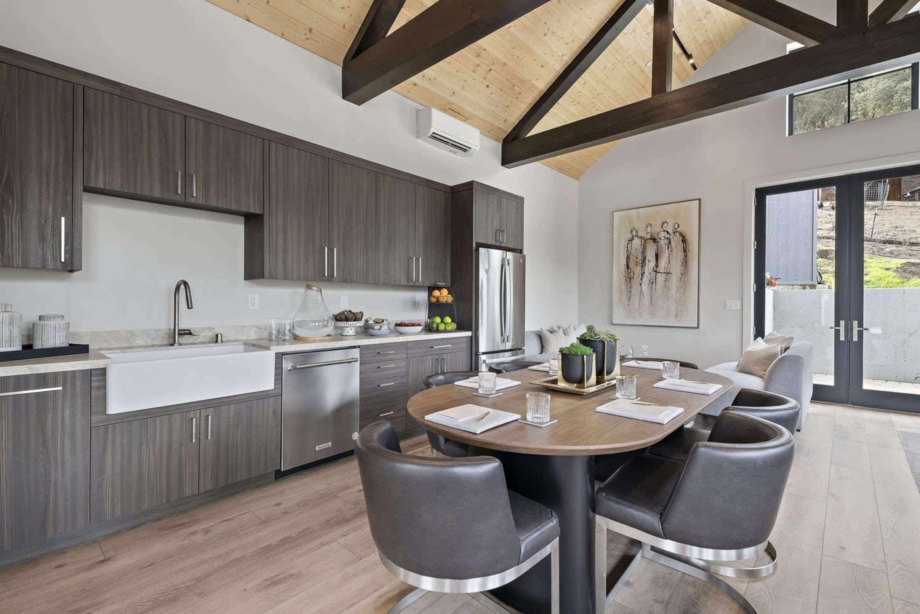 The image shows a modern kitchen with wooden cabinetry, a central dining table, and large windows allowing natural light to enter.