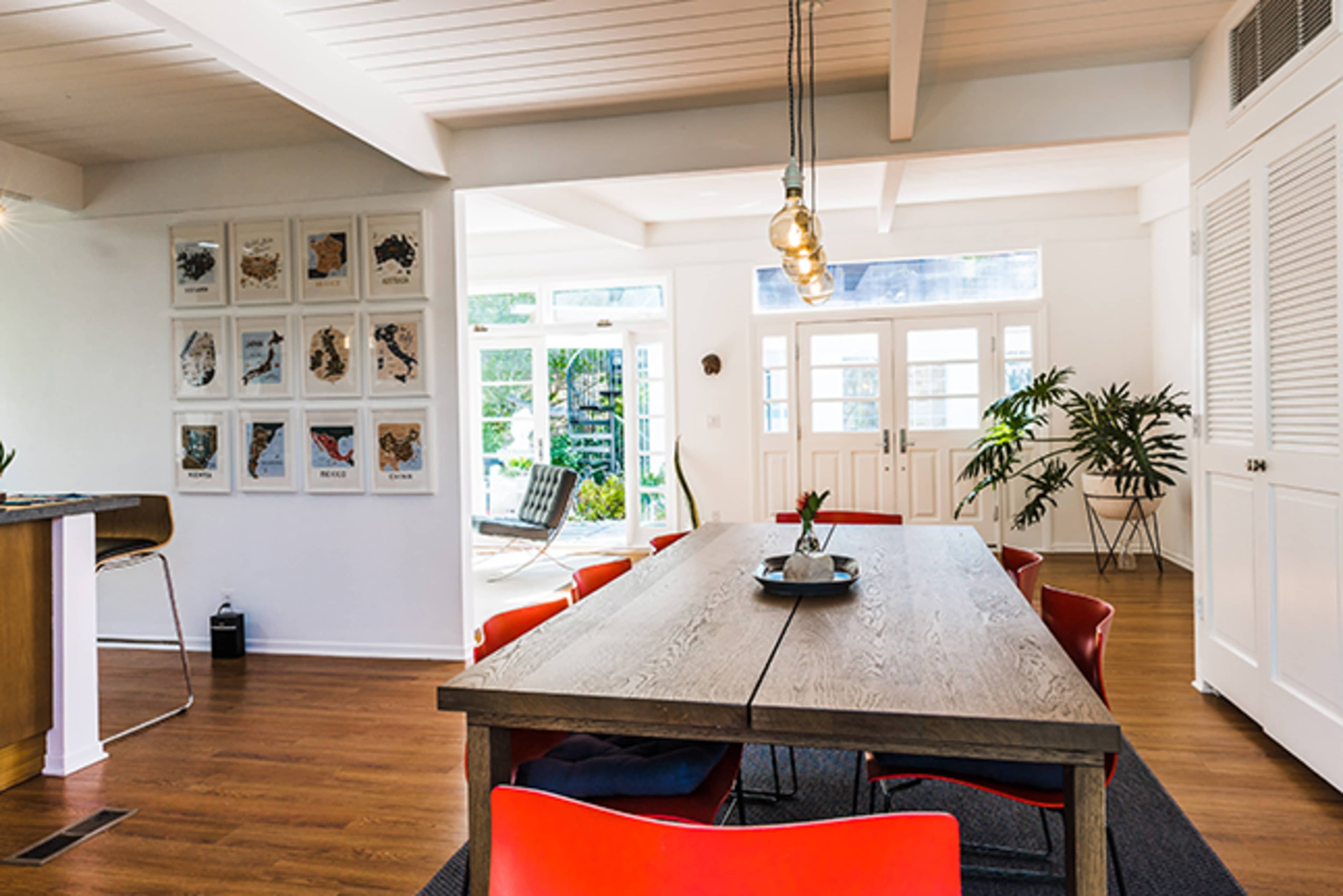 A spacious dining area features a long wooden table surrounded by red chairs and adorned with wall art, leading to a light-filled room with large windows and greenery.