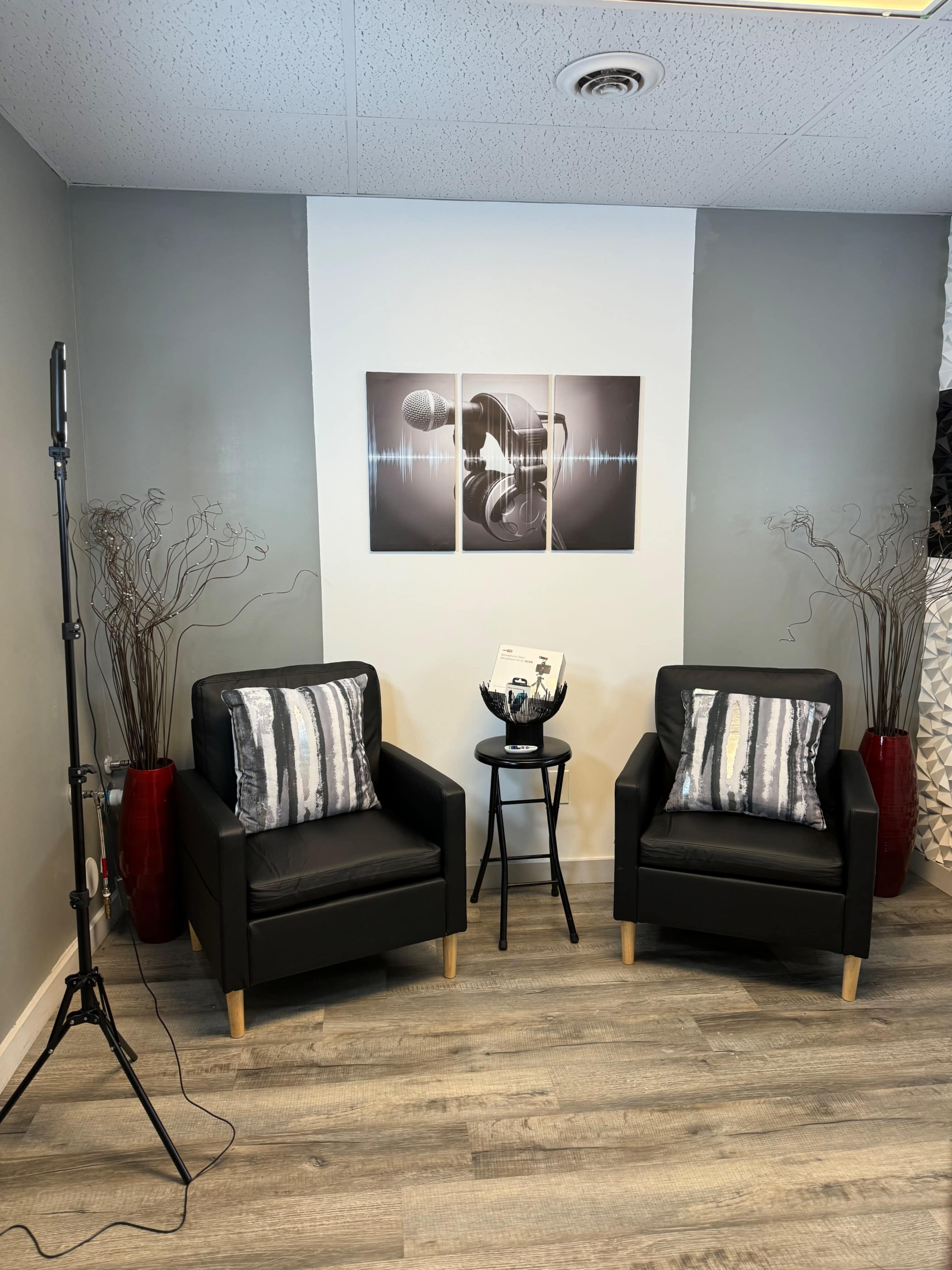 The image shows a cozy corner with two black chairs, a small black table, and decorative artwork on the wall, surrounded by potted plants.