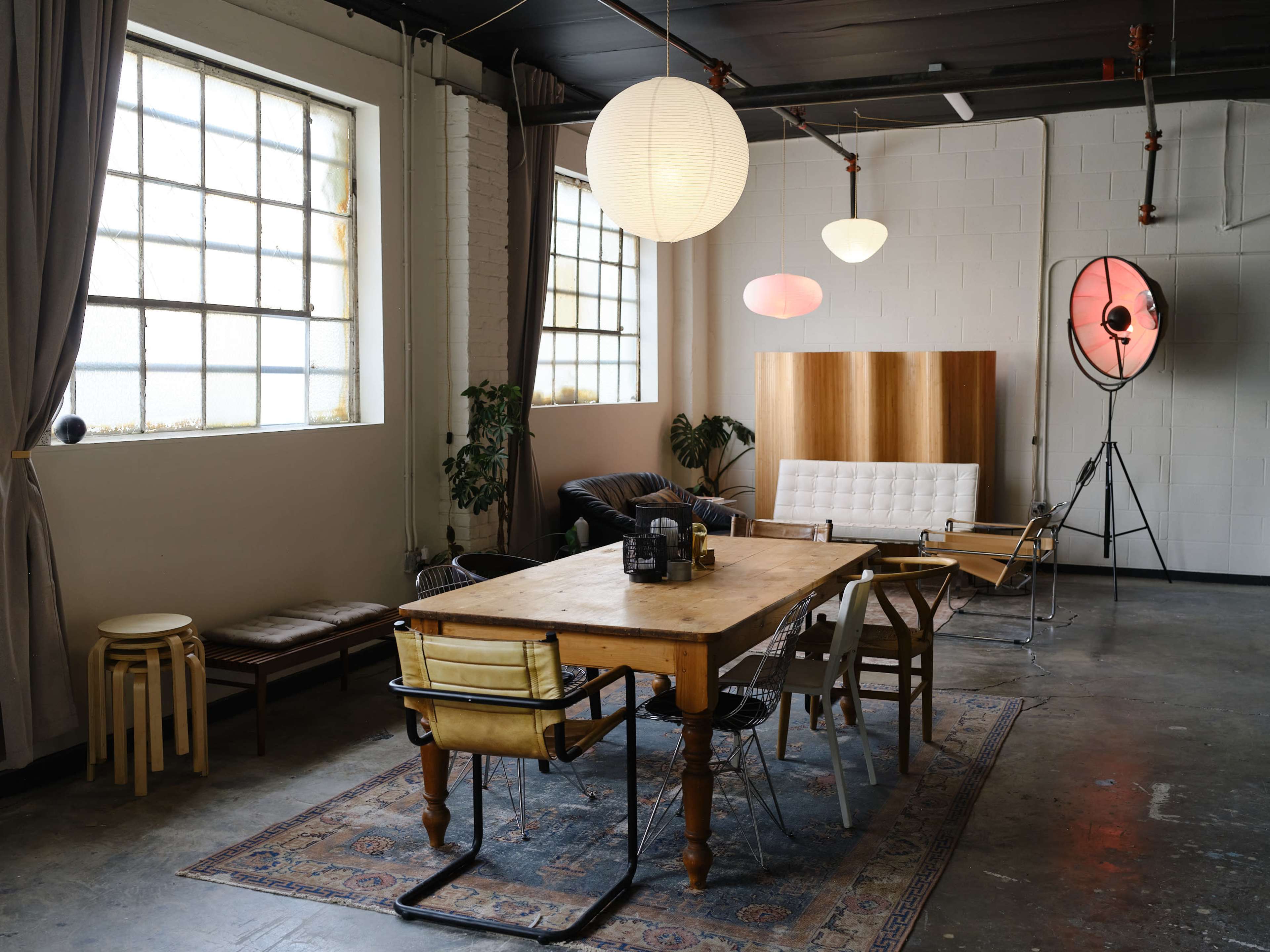A spacious interior featuring a wooden dining table surrounded by various chairs, large windows, and soft lighting fixtures.