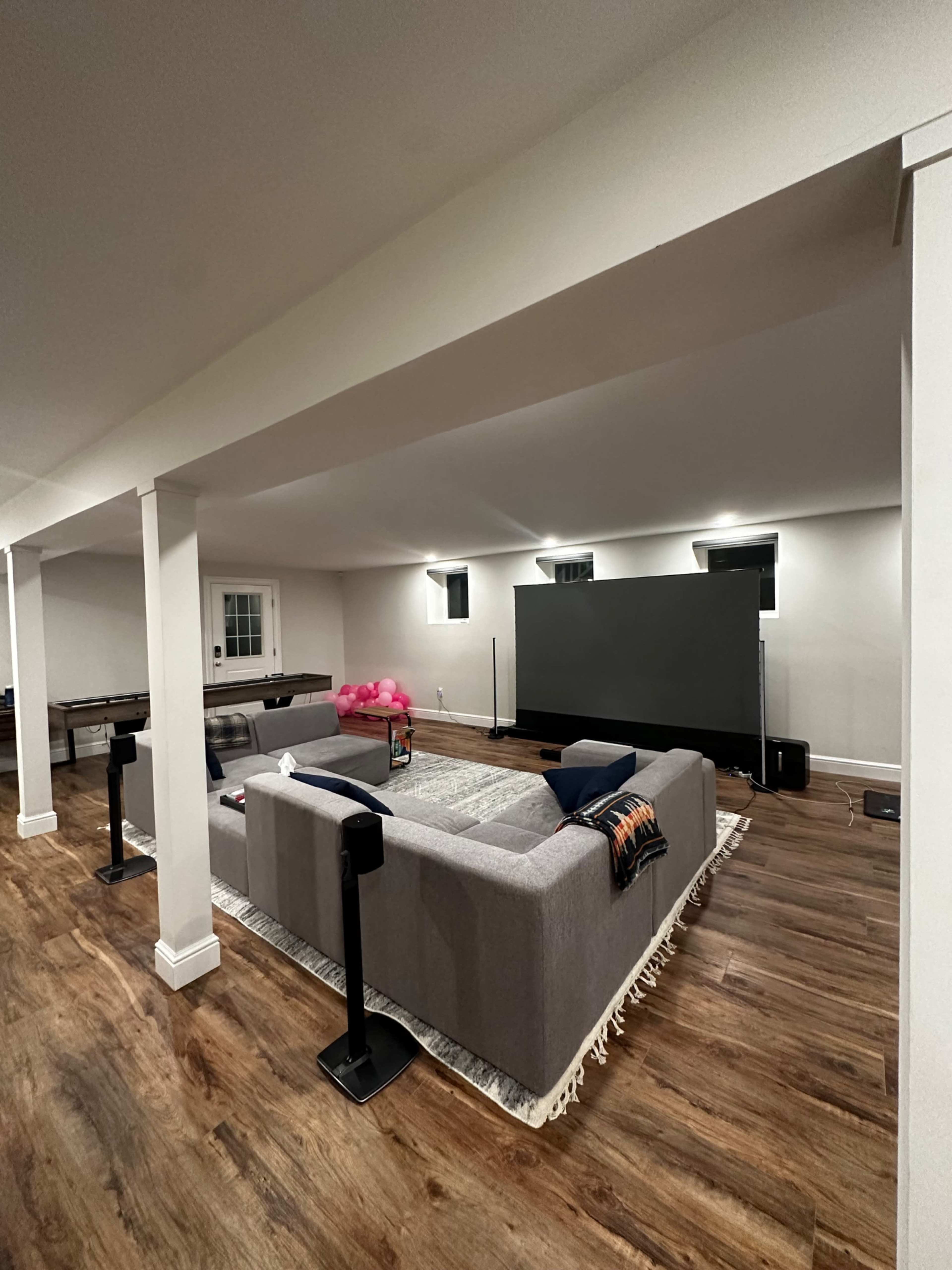 A spacious basement living area features a large gray sectional sofa facing a wall-mounted television, with wooden flooring and lit windows.