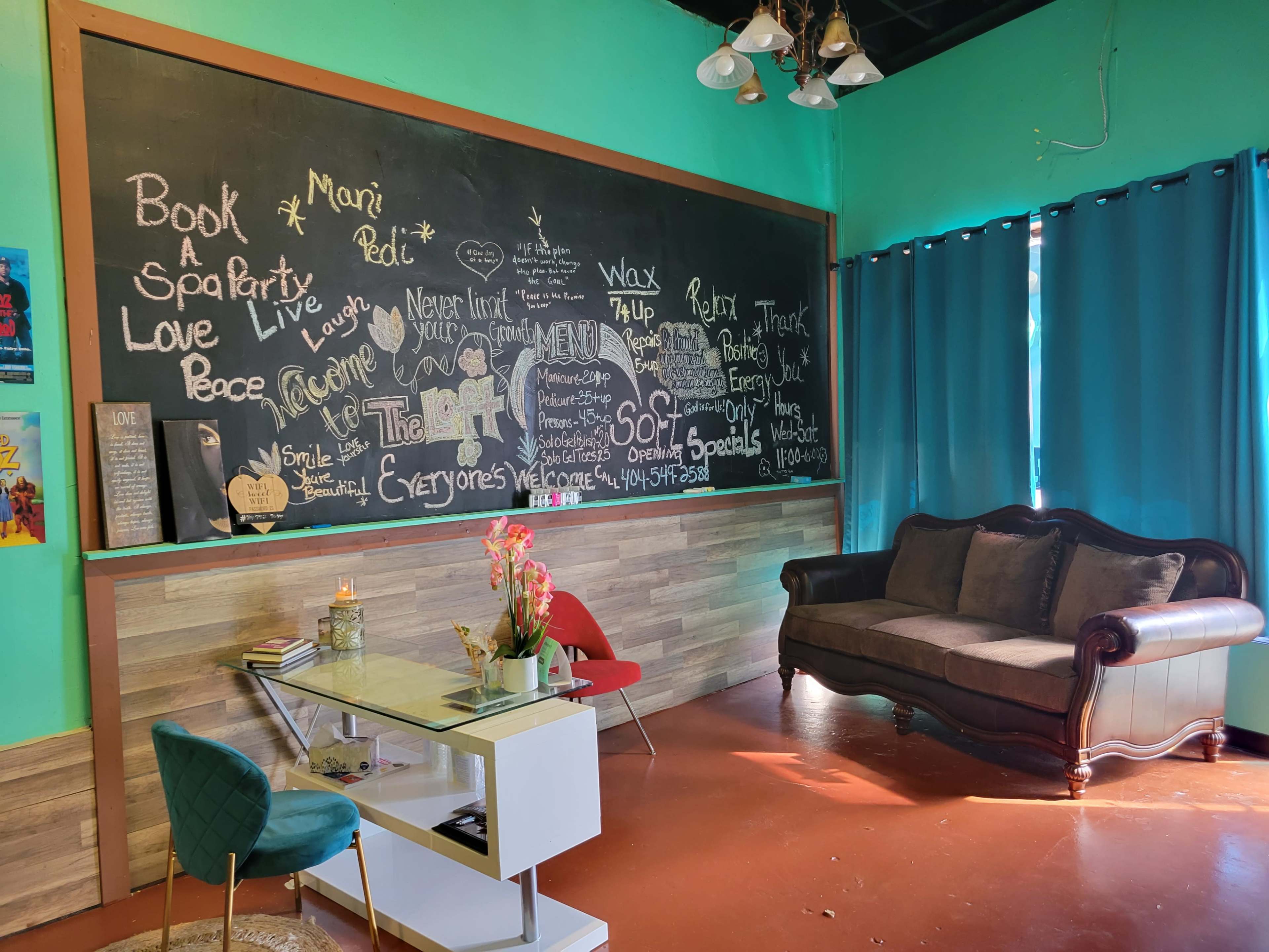 A cozy lounge area with a black chalkboard wall filled with writing, a stylish sofa, a small table, and decorative plants, all set against a vibrant teal background.