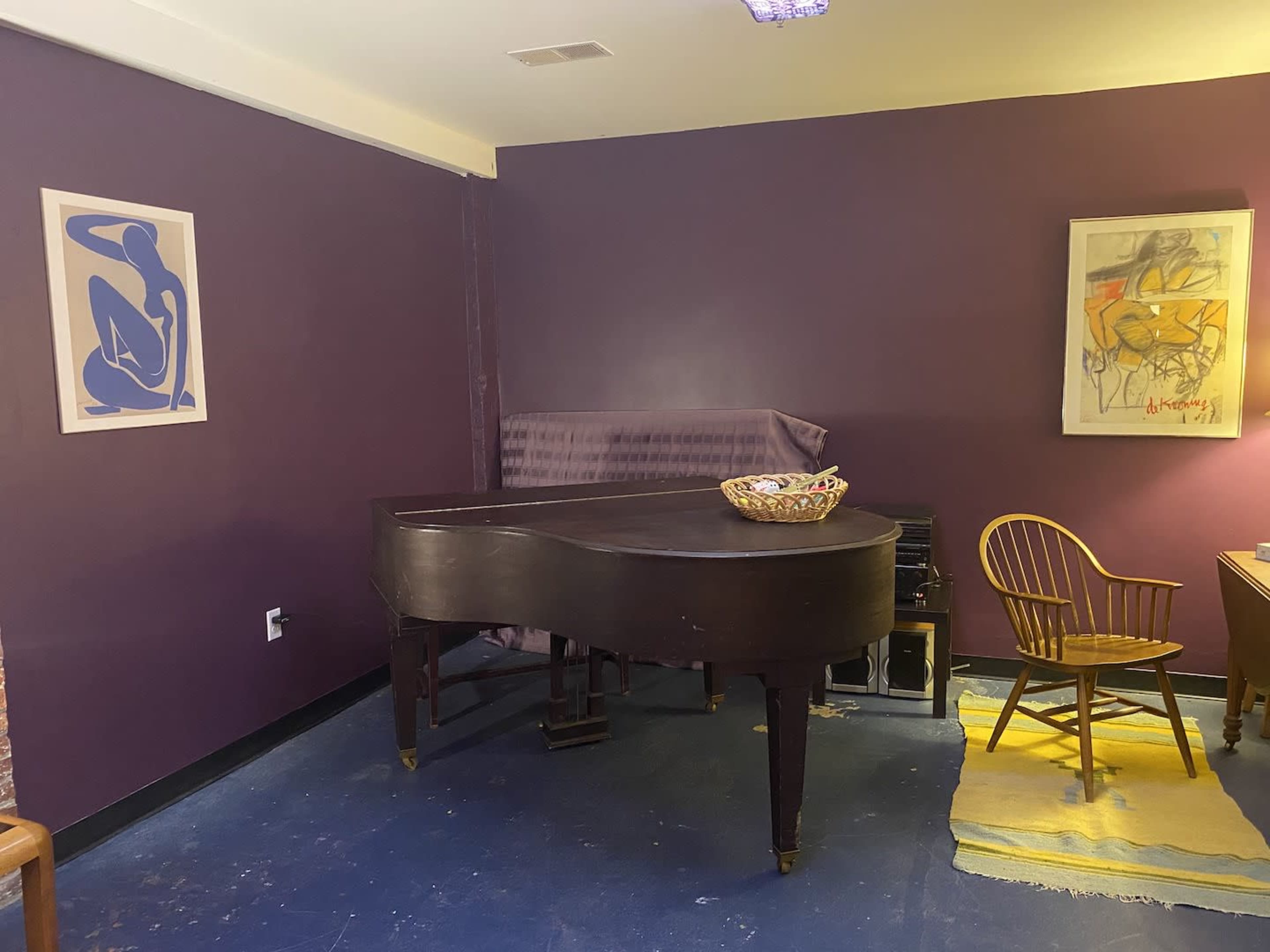 A dimly lit room featuring a grand piano, a wooden chair, and two framed artworks on purple walls.