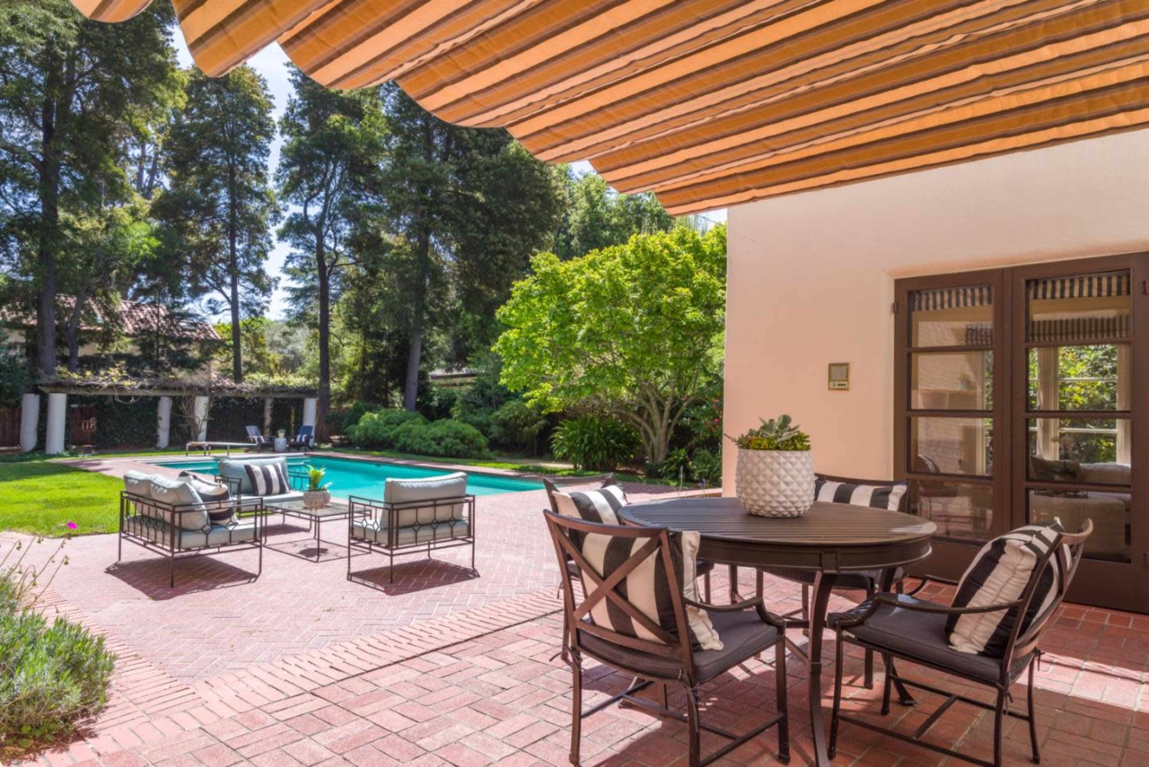 A brick patio with a dining table and chairs overlooks a pool surrounded by trees and landscaped greenery.