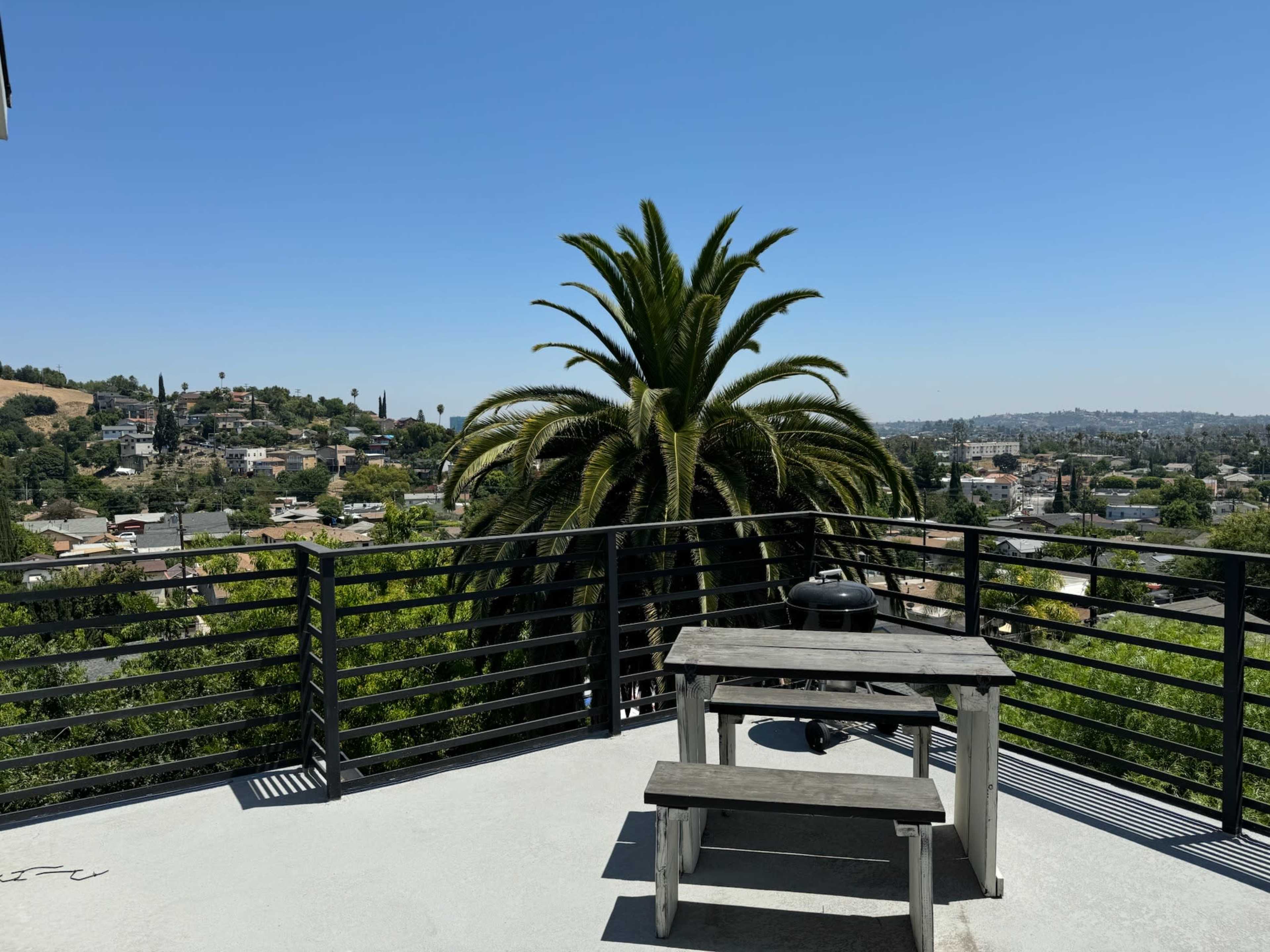 A wooden picnic table sits on a terrace surrounded by a railing, with a large palm tree and a neighborhood view in the background under a clear blue sky.