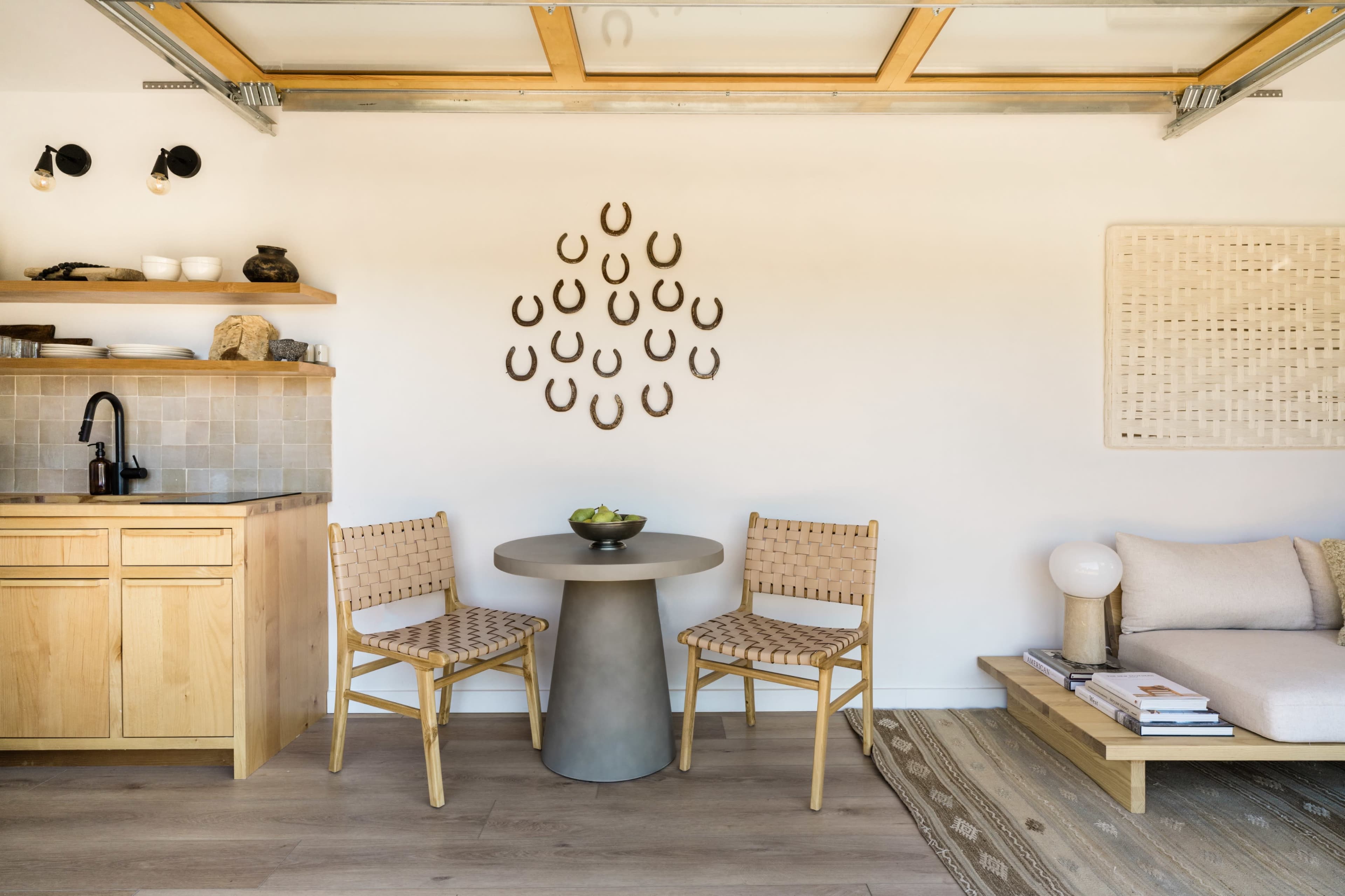 A minimalist kitchen area features wooden cabinets, a round table with two woven chairs, and a cozy seating space, accented by a wall display of horseshoes.