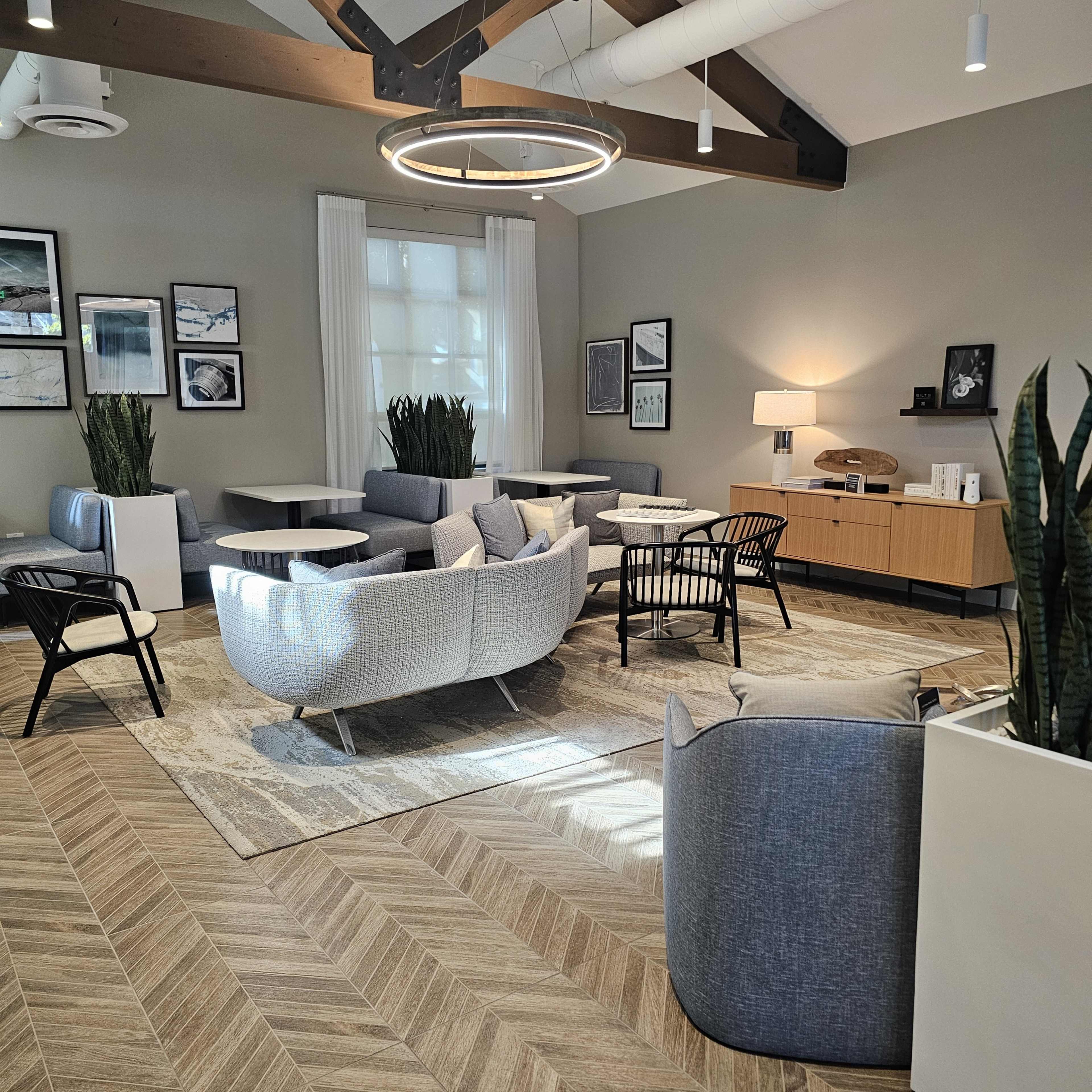 A modern indoor lounge area with comfortable seating, a wooden coffee table, and decorative plants, illuminated by a circular hanging light fixture.