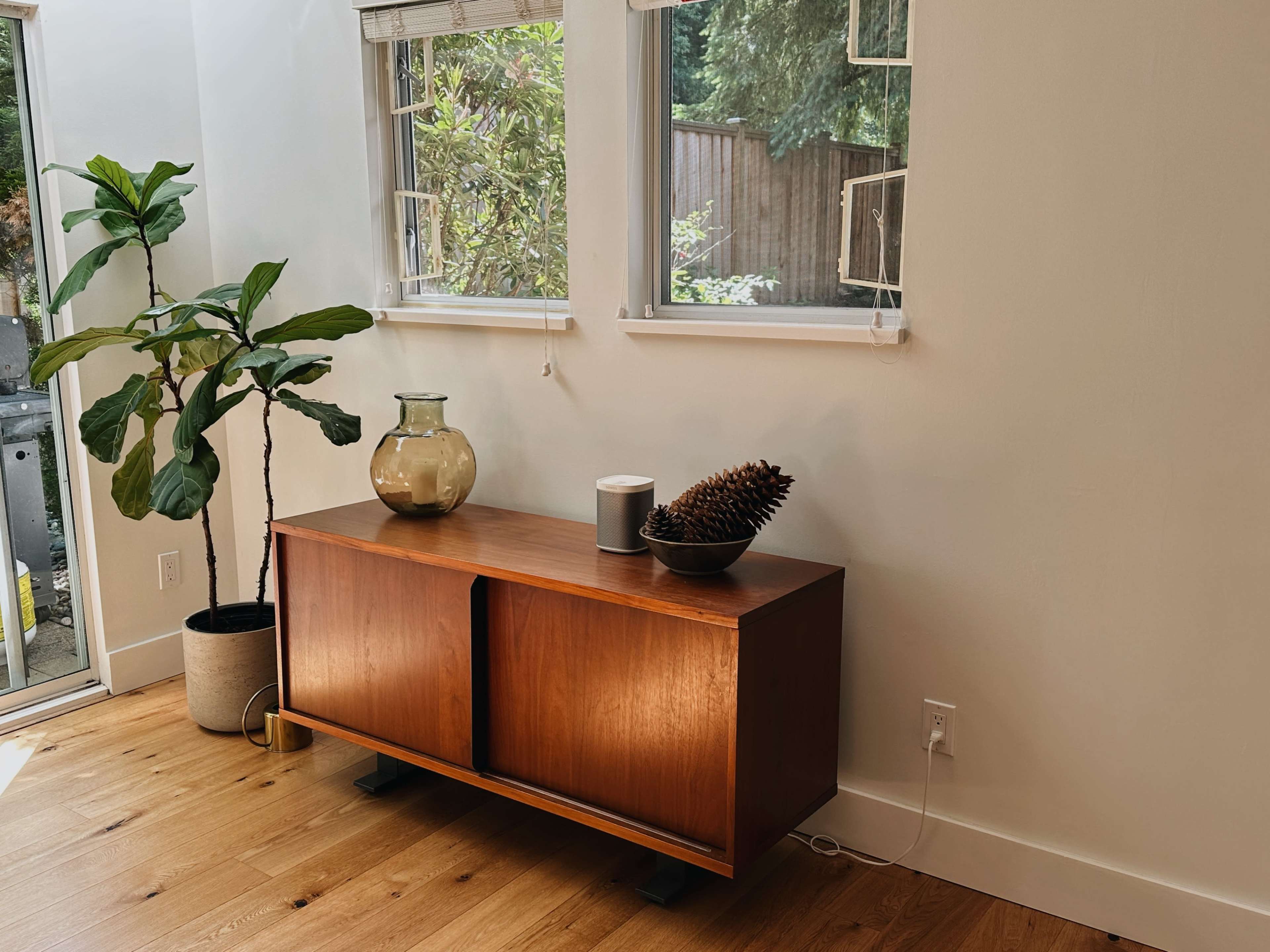The image features a wooden sideboard with sliding doors, accompanied by a glass vase, a candle, and a pinecone, set against a backdrop of large windows and a potted plant.