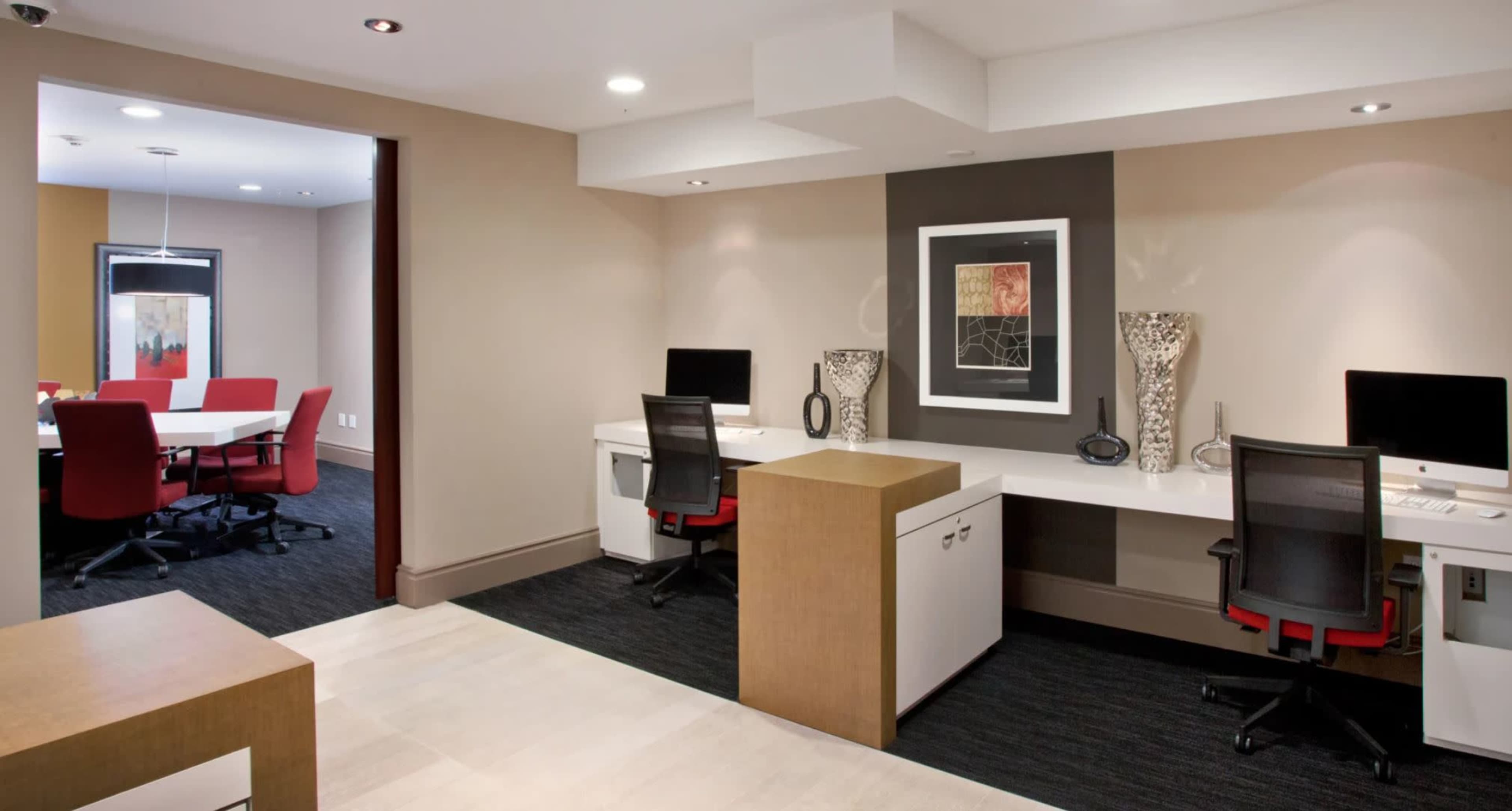 The image shows an office space featuring two workstations with computers and a meeting room in the background with red chairs around a table.