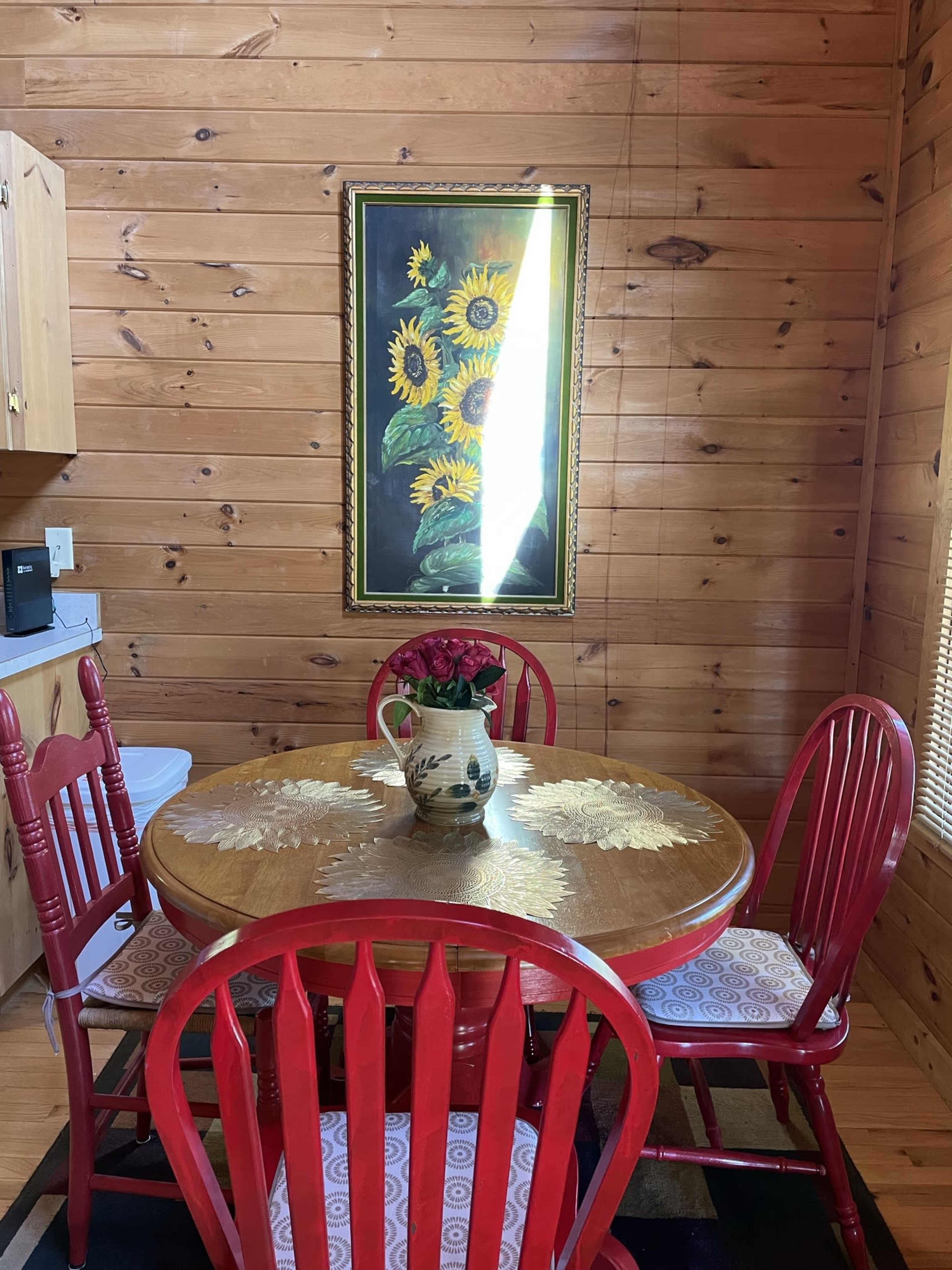 A round wooden table with red chairs is set in a room featuring wooden walls and a framed painting of sunflowers.