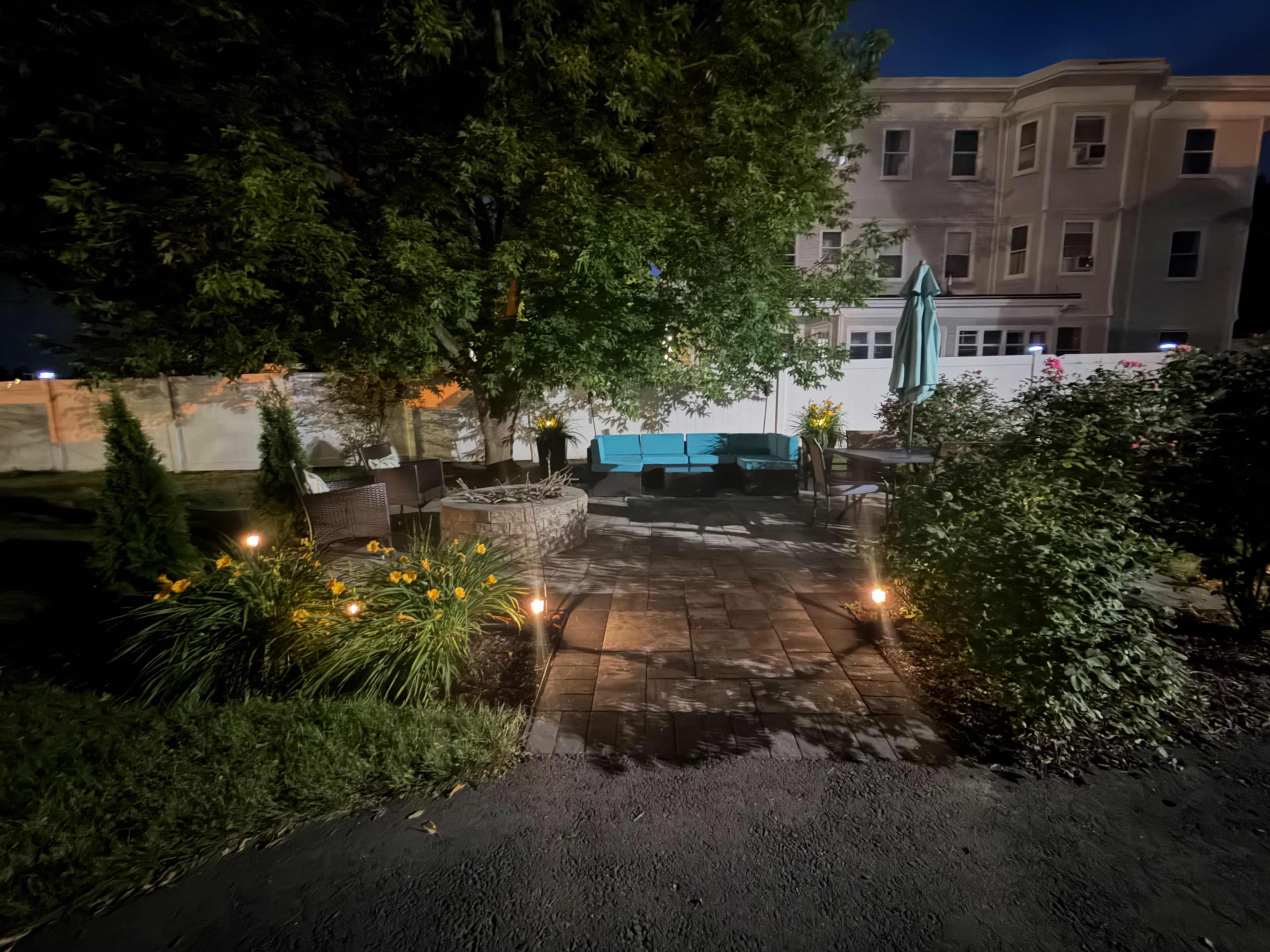 A well-lit patio area features a stone pathway surrounded by greenery and seating, with a large tree providing shade and a building visible in the background.