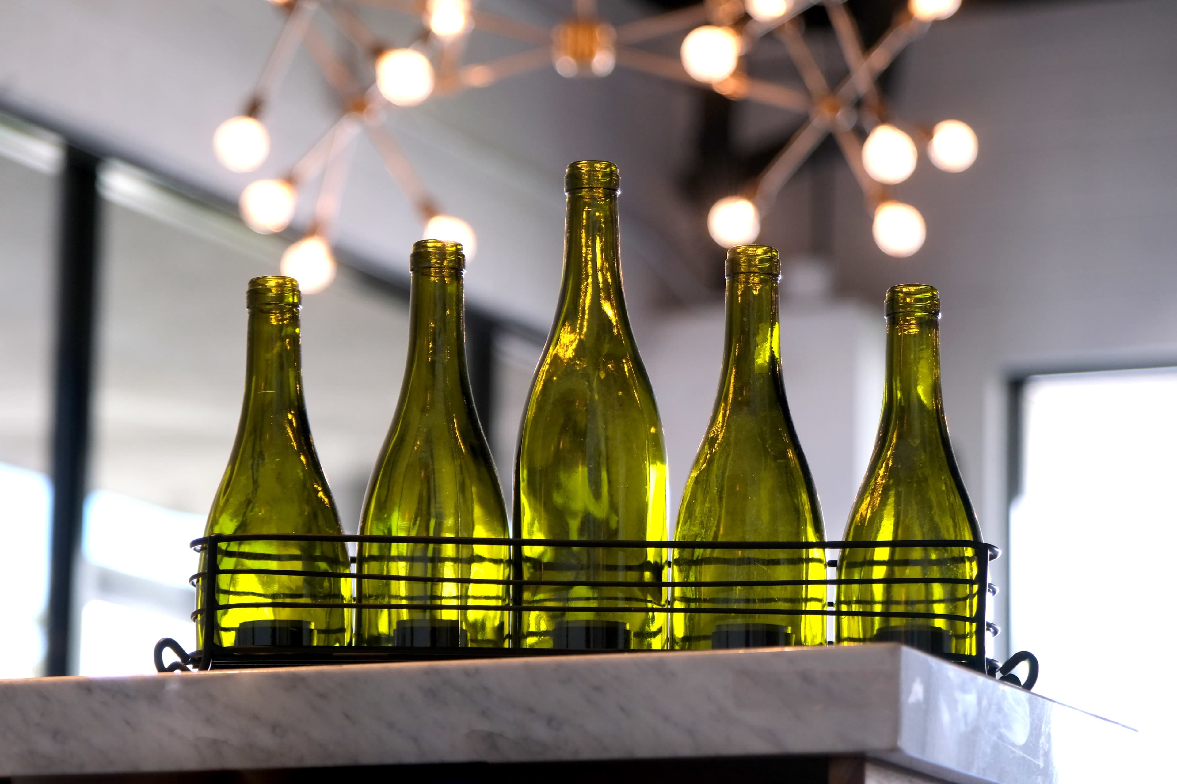 Five green glass bottles are arranged in a wire basket on a marble surface, with a light fixture visible in the background.
