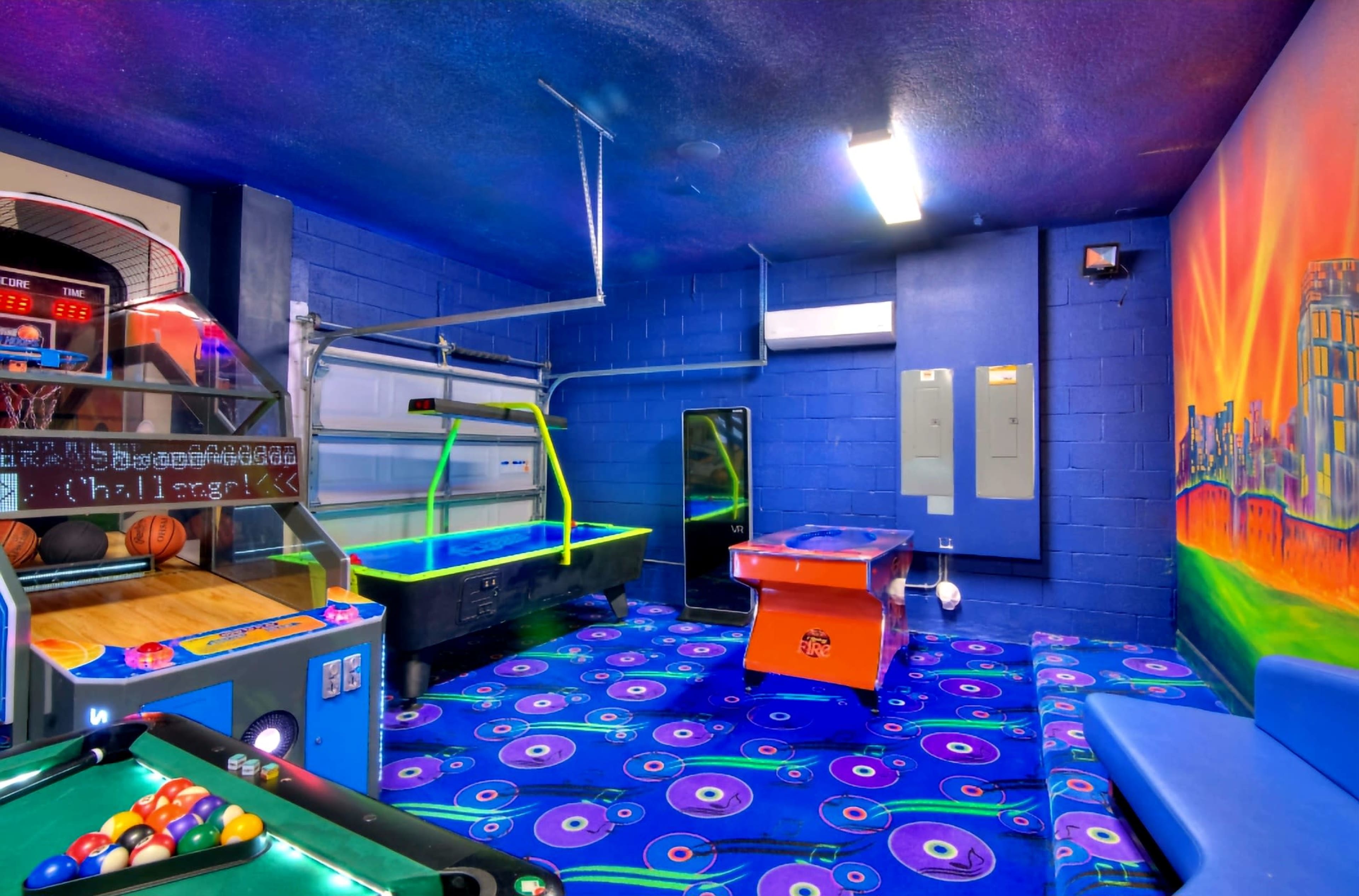 A vibrant game room equipped with a pool table, air hockey table, and a basketball arcade game, all set against a blue wall and patterned carpet.