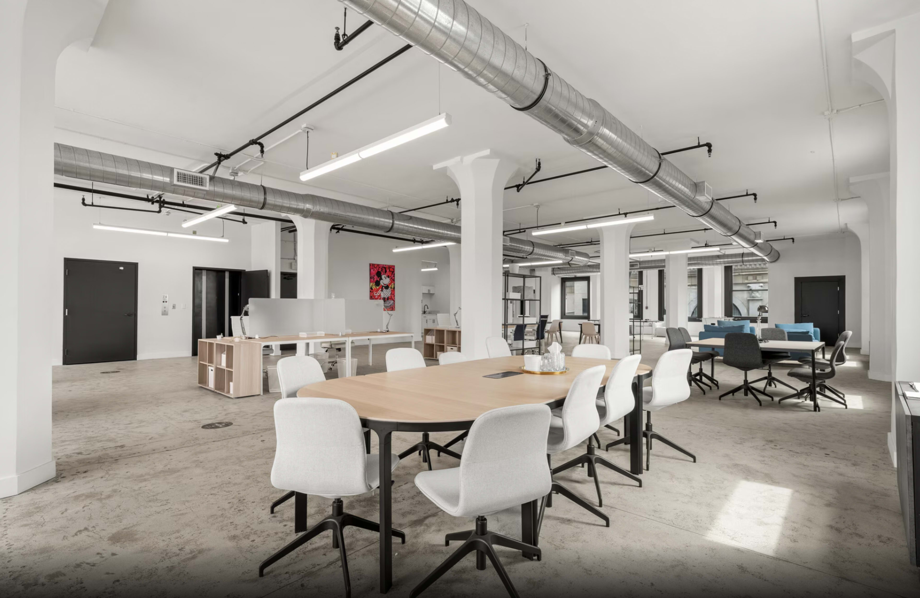 The image shows a modern office space featuring multiple tables and chairs, large windows, and industrial-style ductwork exposed on the ceiling.