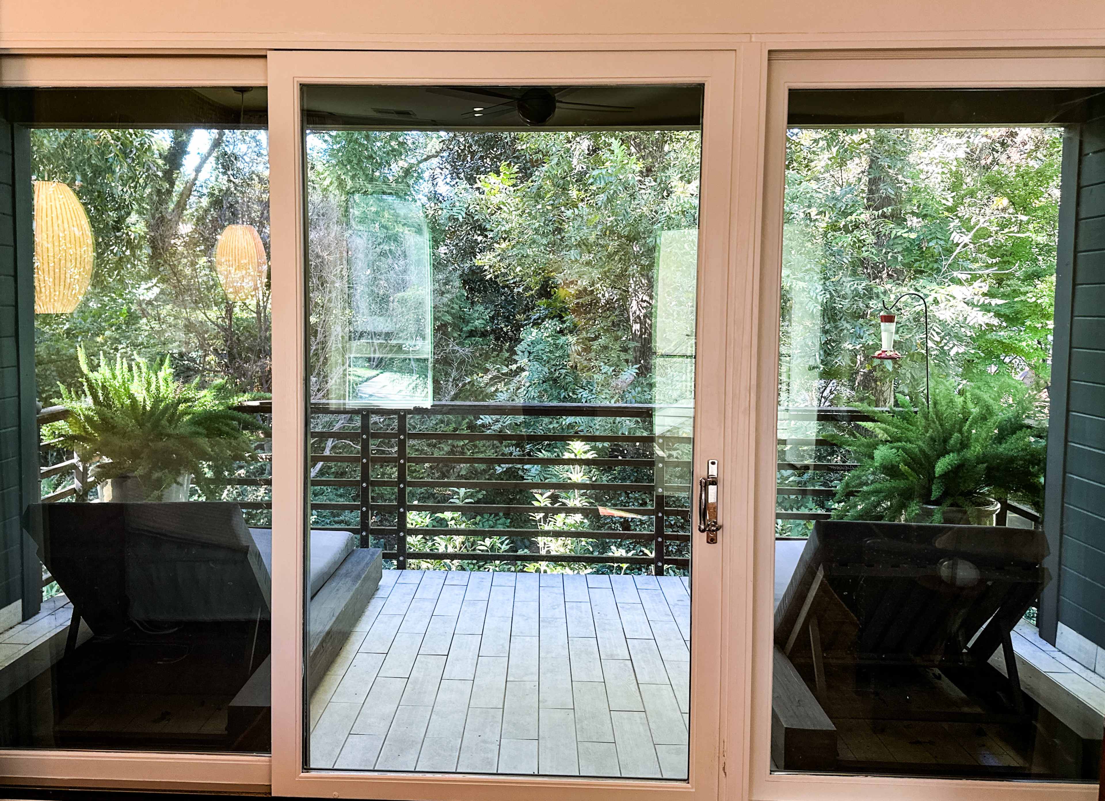 The image shows a glass door leading to a wooden balcony surrounded by greenery, with two lounge chairs and potted ferns on each side.