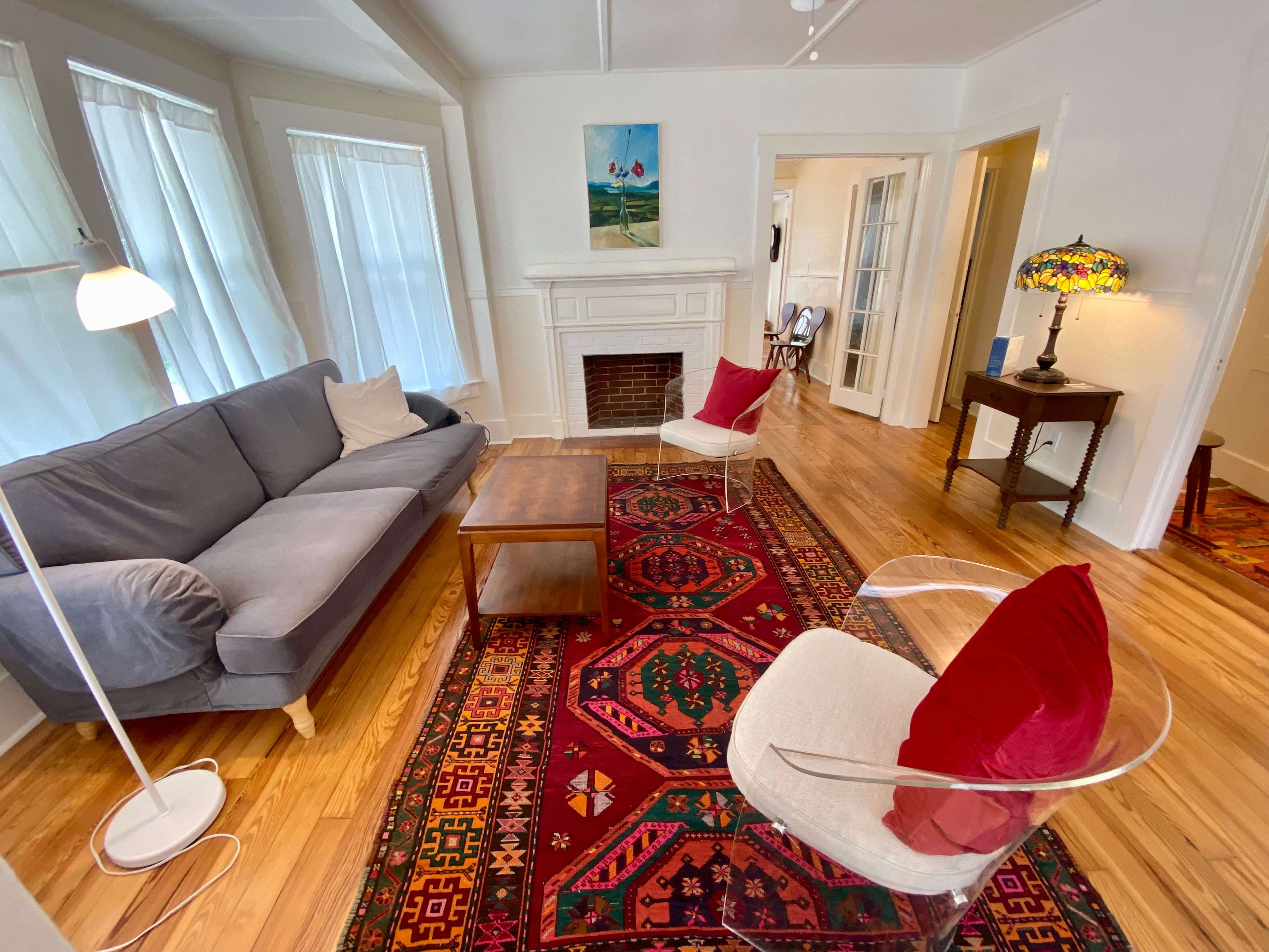 The living room features a gray sofa, two white chairs with red cushions, a wooden coffee table, and a colorful rug, along with a fireplace and artwork on the walls.