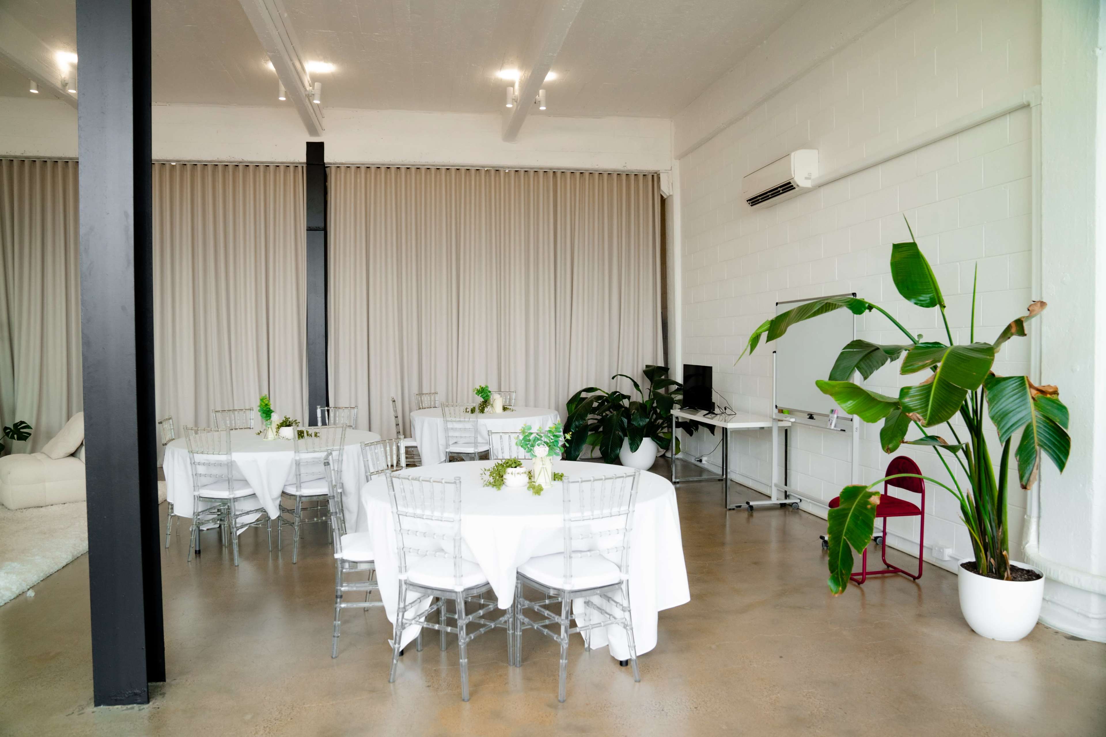 The image shows a modern event space with white tables set for a gathering, featuring green floral centerpieces and large potted plants beside a wall of beige curtains.