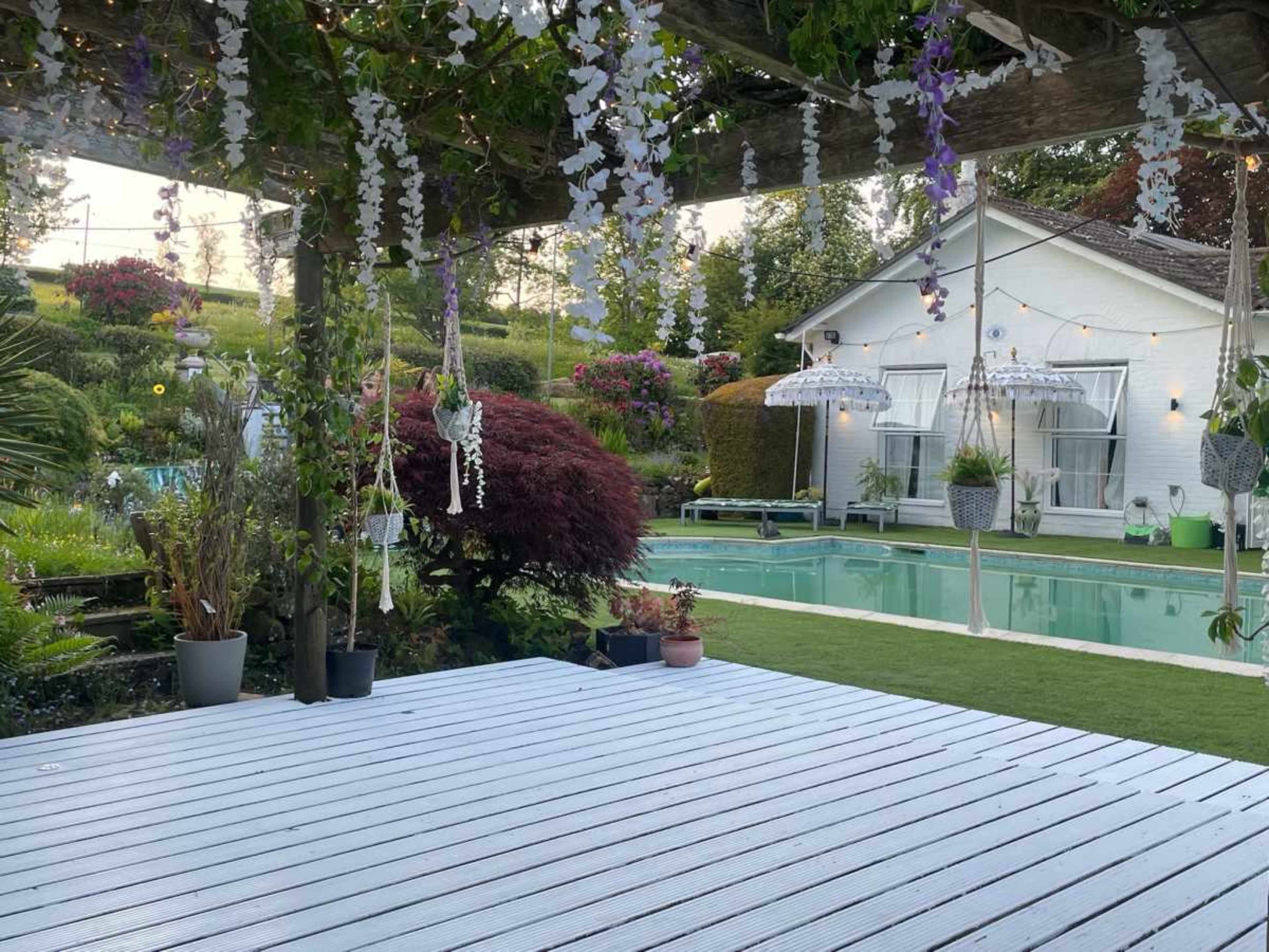 A serene backyard with a swimming pool, lush greenery, and a white house in the background, framed by hanging plants and decorative elements.