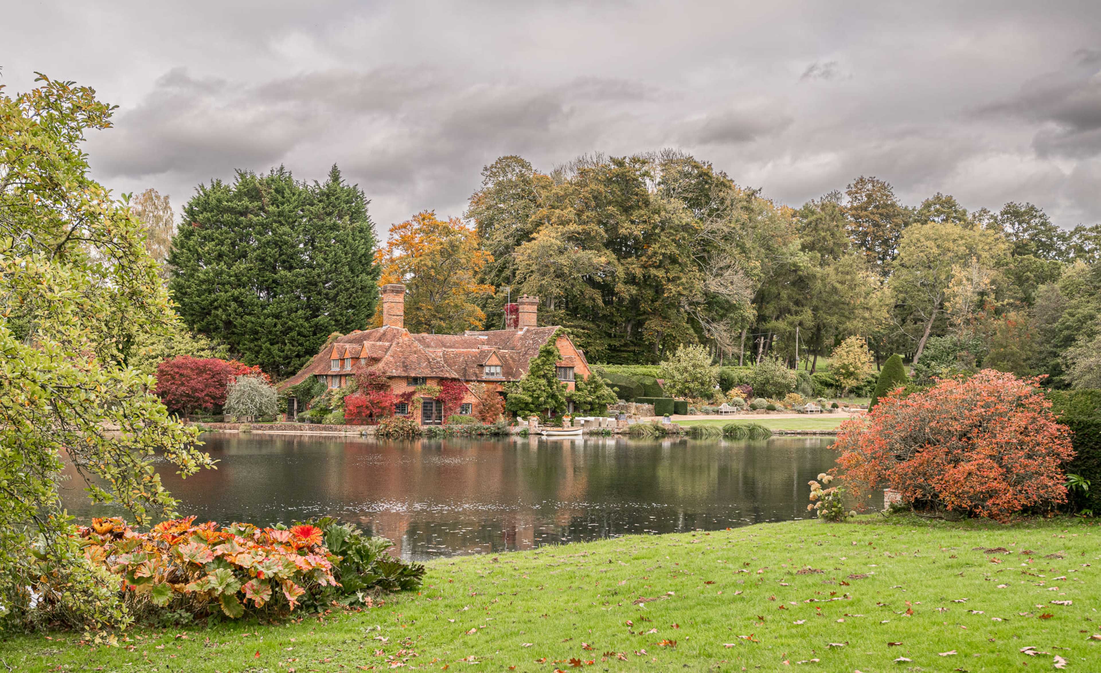 A charming house with a thatched roof sits beside a still lake, surrounded by trees displaying autumn colors.