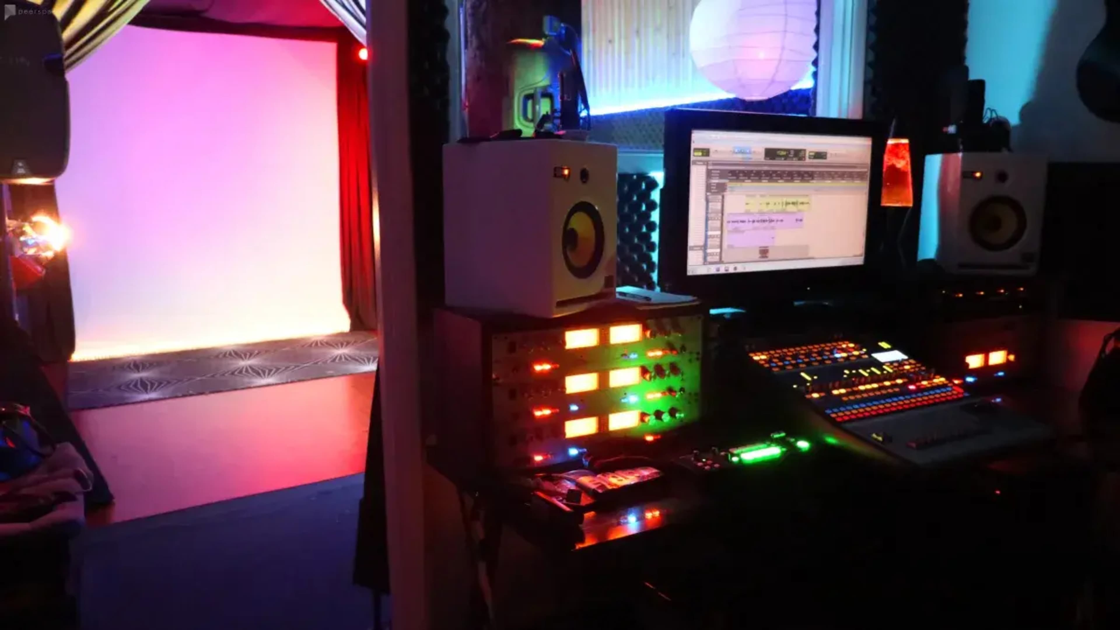 A music production studio equipped with a computer, audio mixing console, and colorful LED lighting, leading to a room with a blank backdrop.