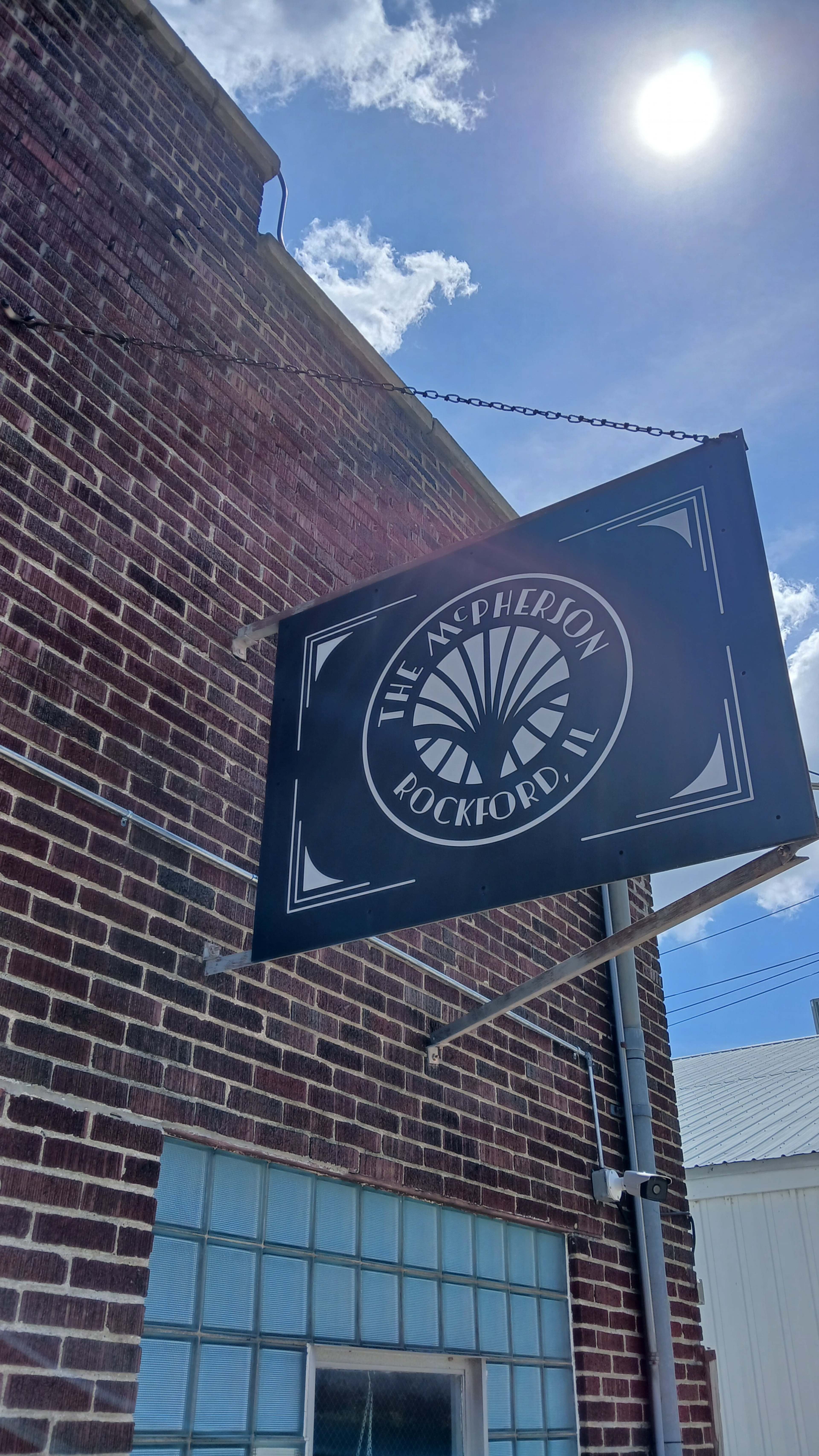 A black sign with a circular design and the text "THE McPHERSON ROCKFORD, IL" hangs from a brick building under a bright sun.
