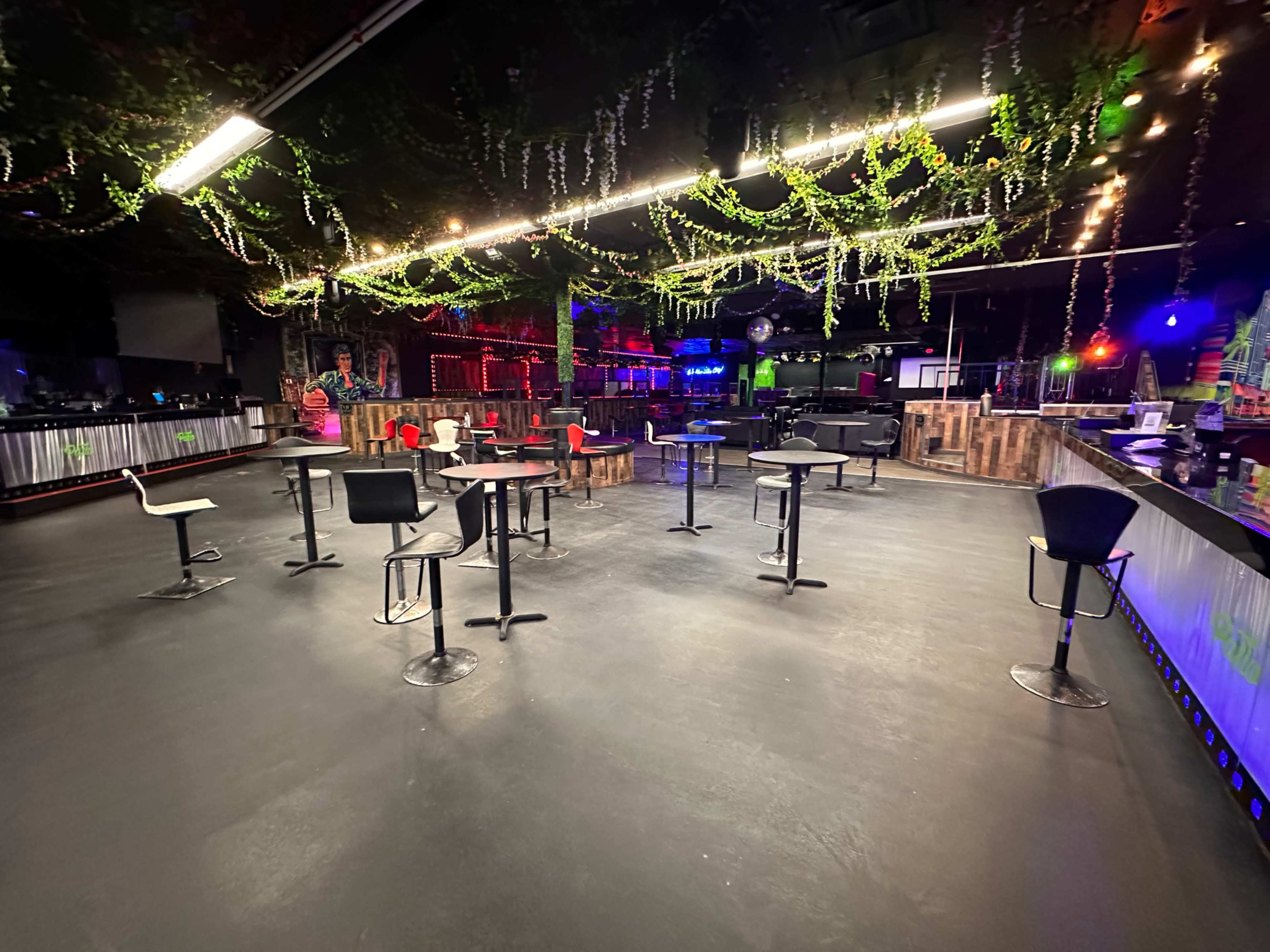 An empty bar area with scattered high tables and chairs under dim, colorful lighting.