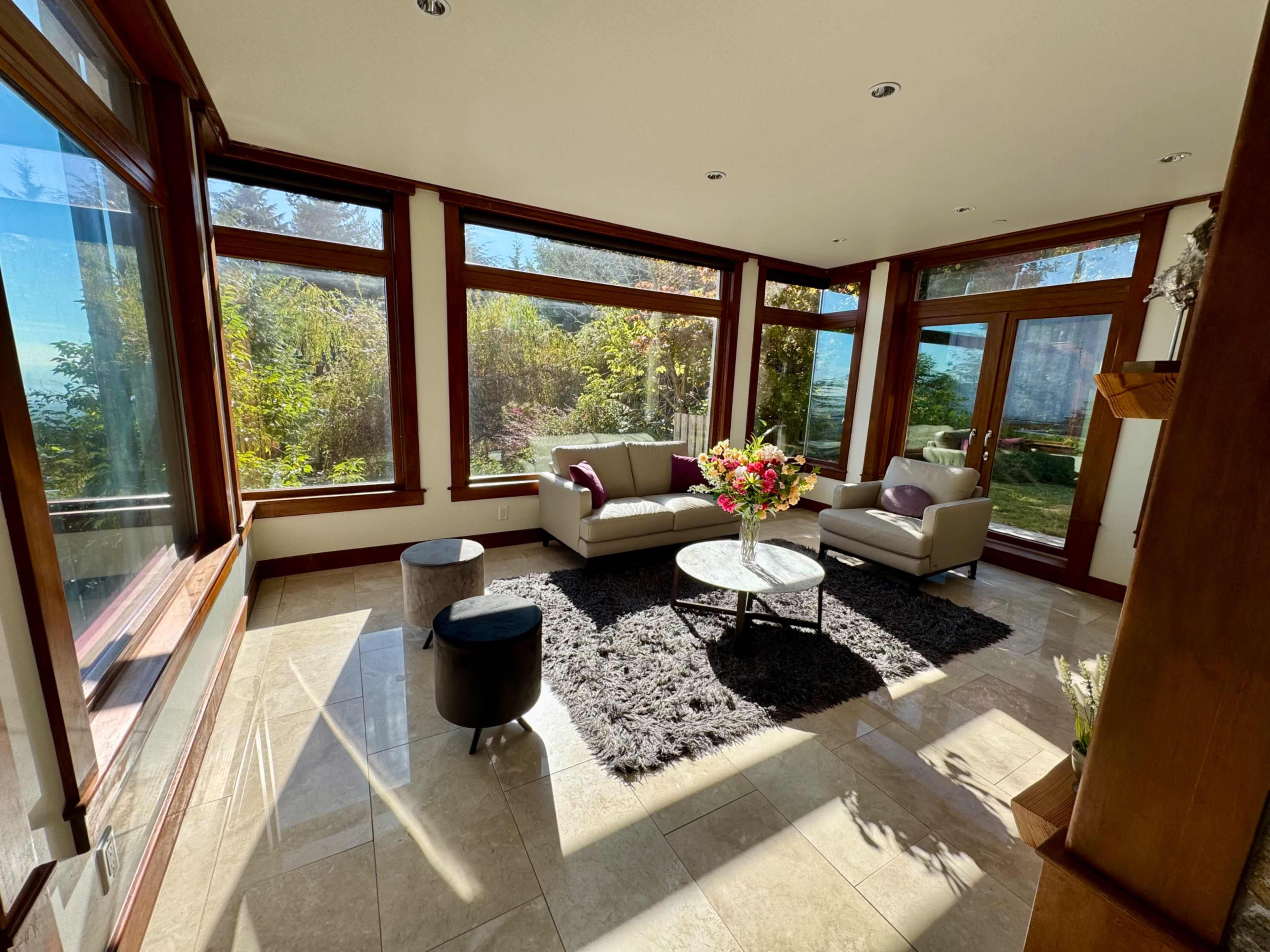 The image shows a sunlit living space with large windows, a beige sofa, a round coffee table, and a floral centerpiece on a dark rug.