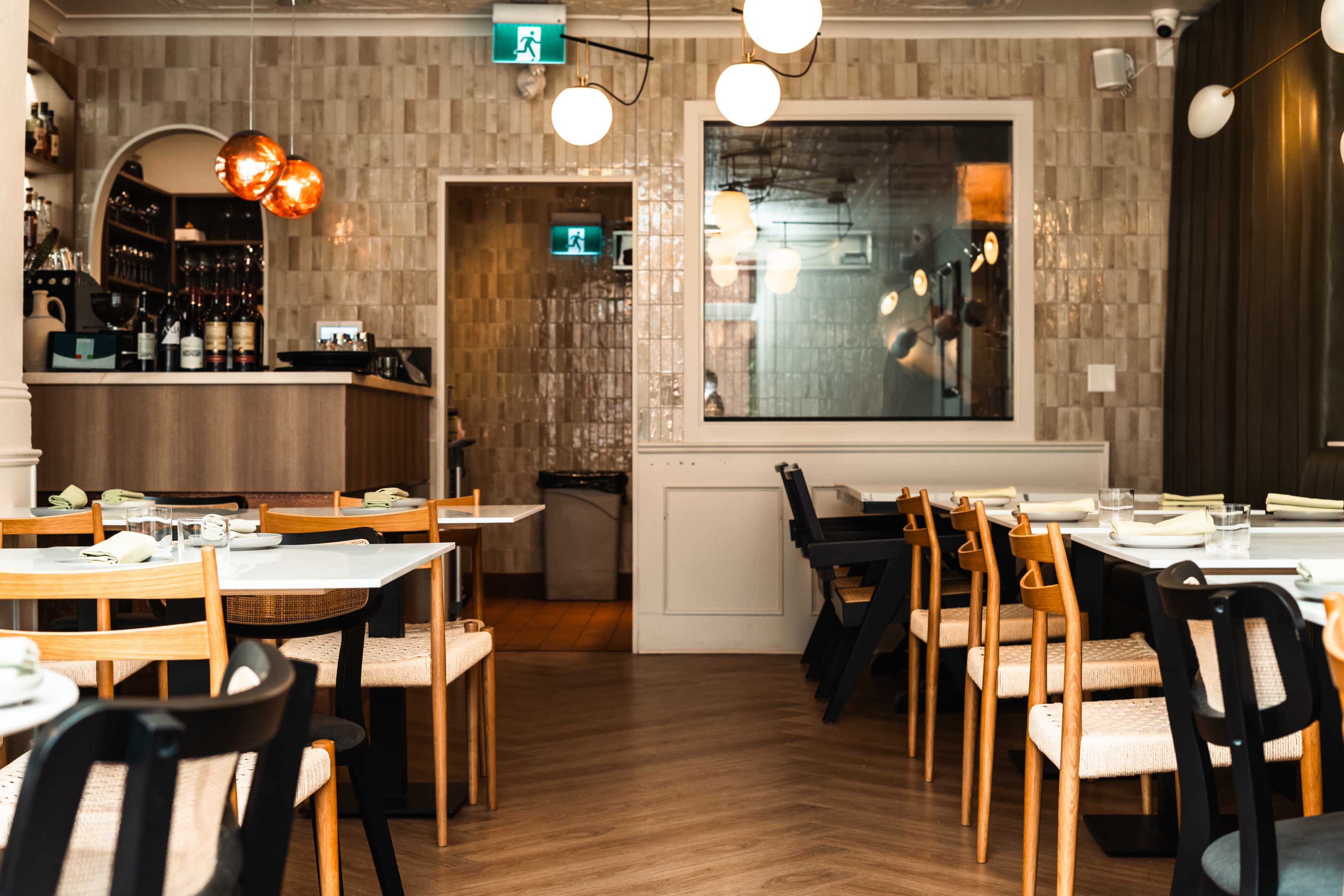 The image shows a modern restaurant interior with wooden tables, assorted chairs, and a bar area in the background.