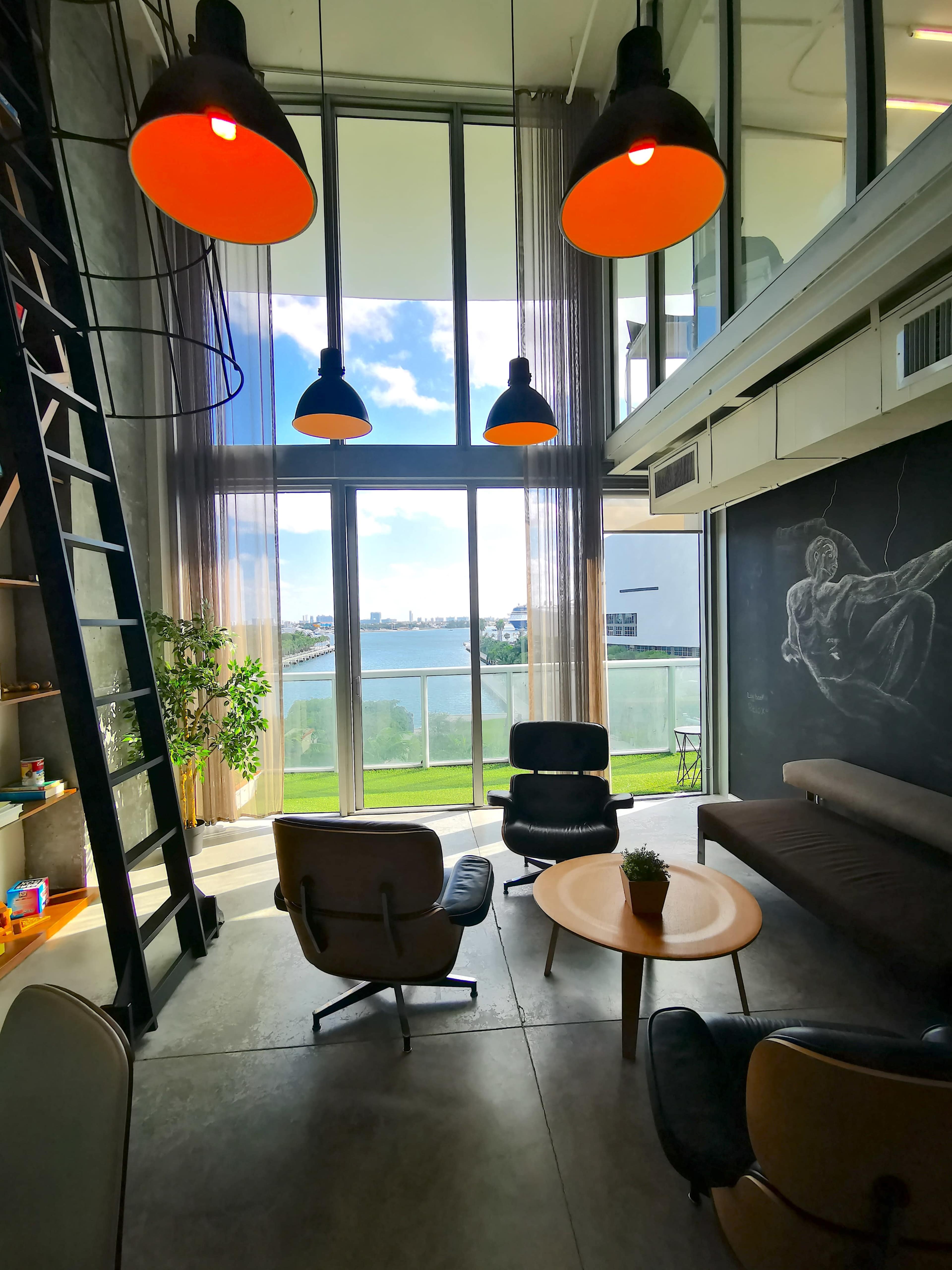 A brightly lit, modern lounge area with large windows overlooking a body of water, featuring stylish furniture and industrial-style lighting.