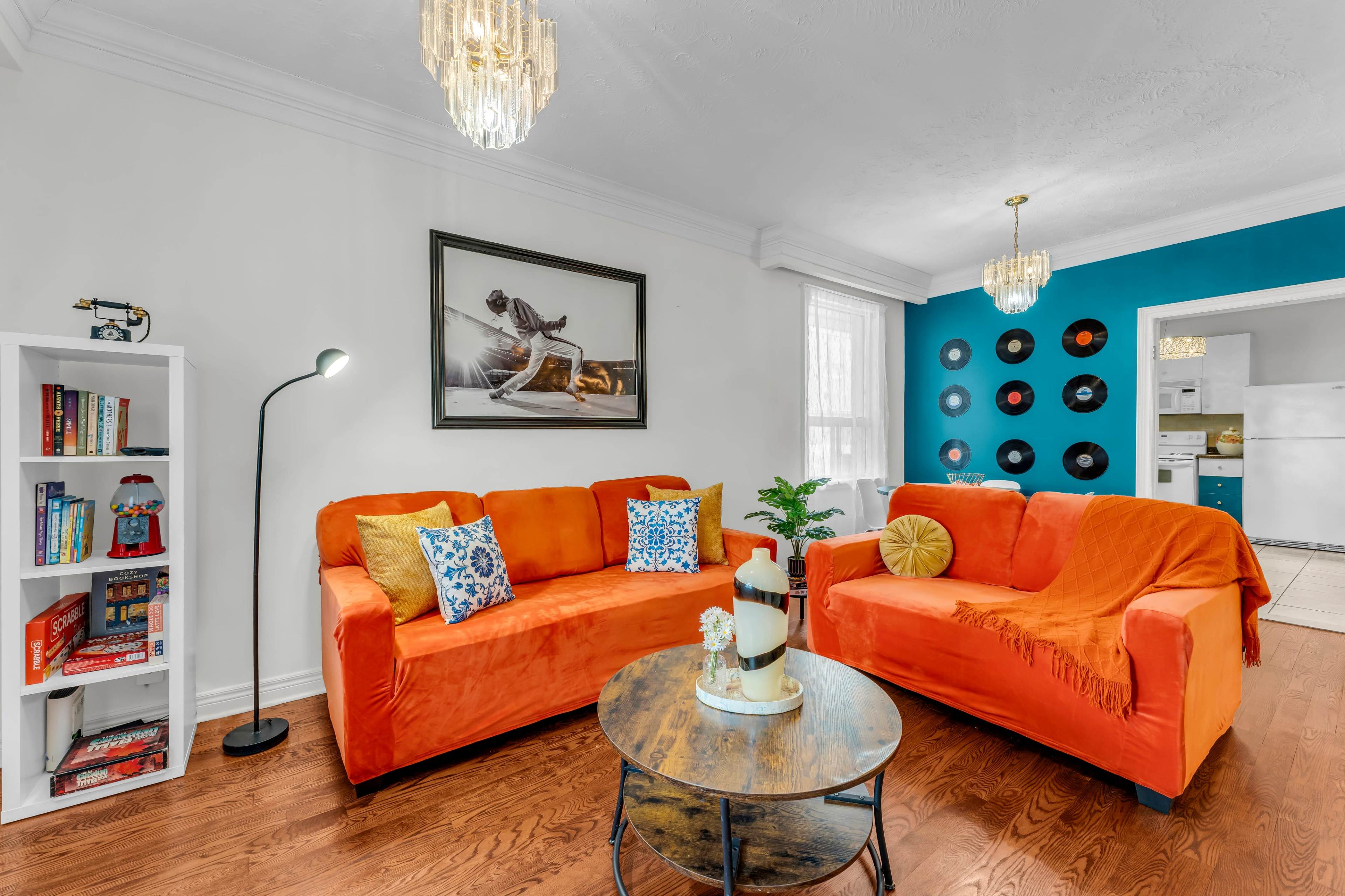 The living room features two orange sofas, a round coffee table, a floor lamp, a bookshelf, and a wall adorned with decorative vinyl records.