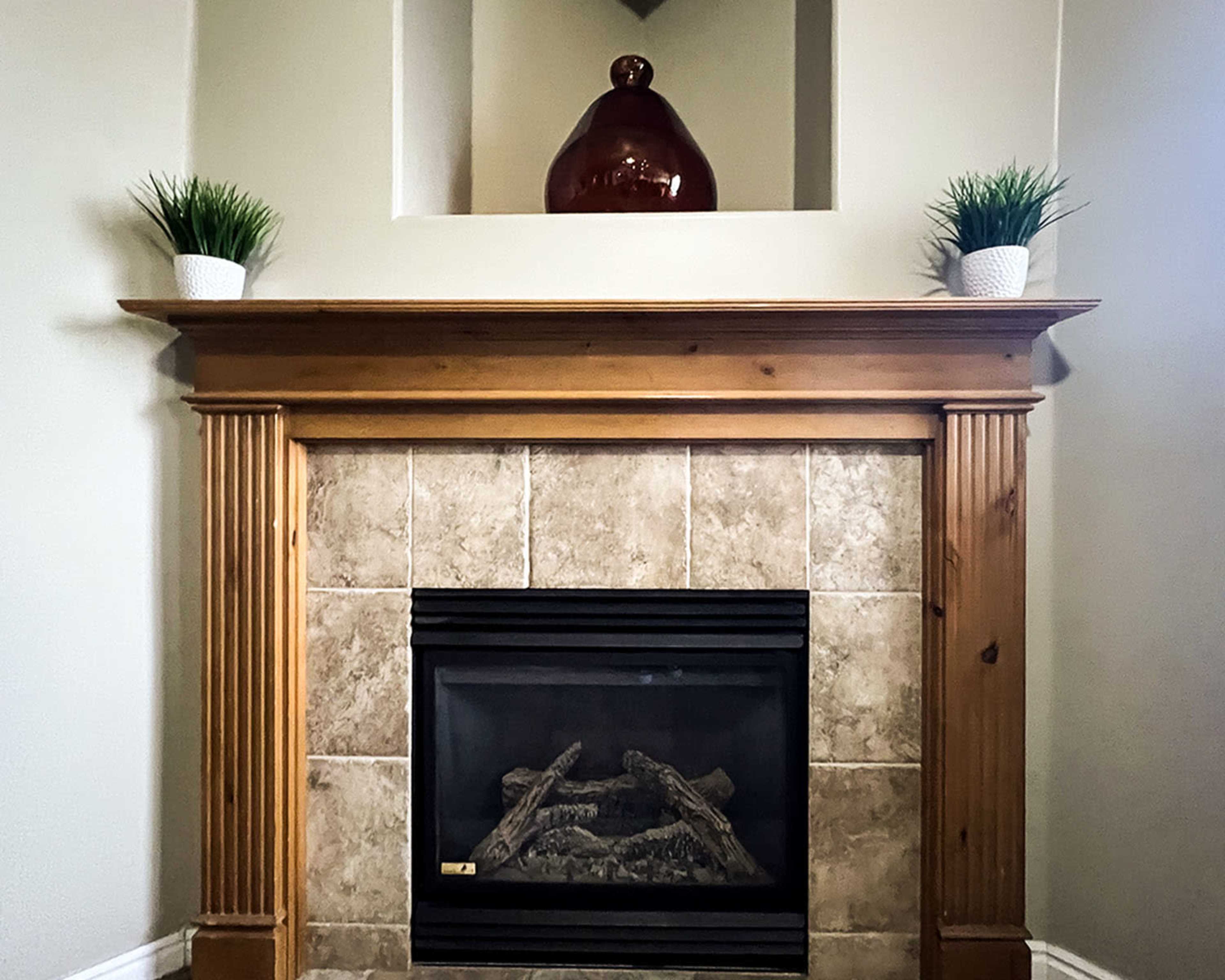 A wooden fireplace mantel topped with two potted plants and a decorative vase above a tiled fireplace.