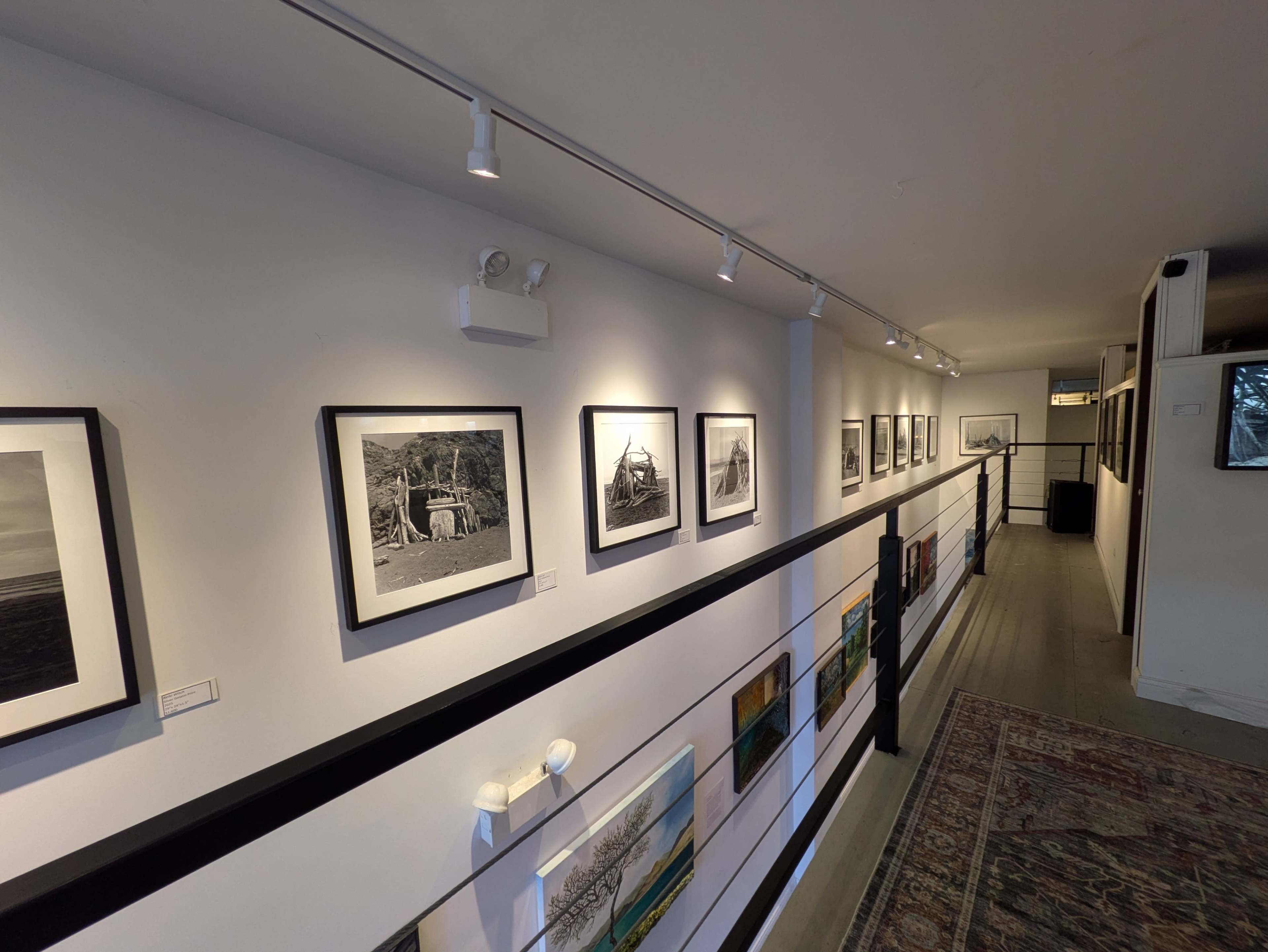 The image depicts an interior gallery with a long white wall featuring a series of framed black-and-white photographs, with additional artwork displayed below them.