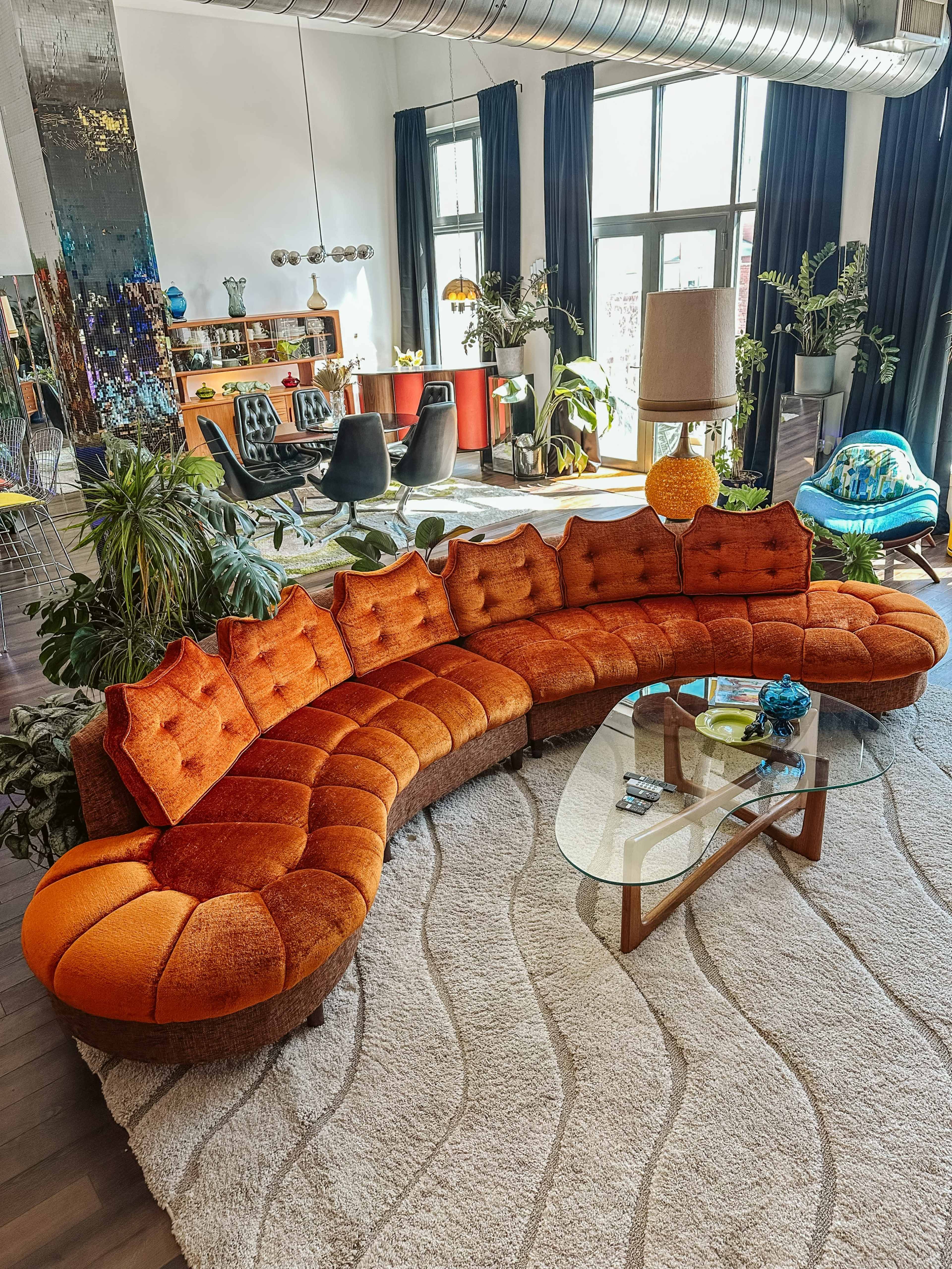 The image features a curved orange velvet sofa arranged around a glass coffee table in a bright, modern living room filled with plants and stylish furniture.