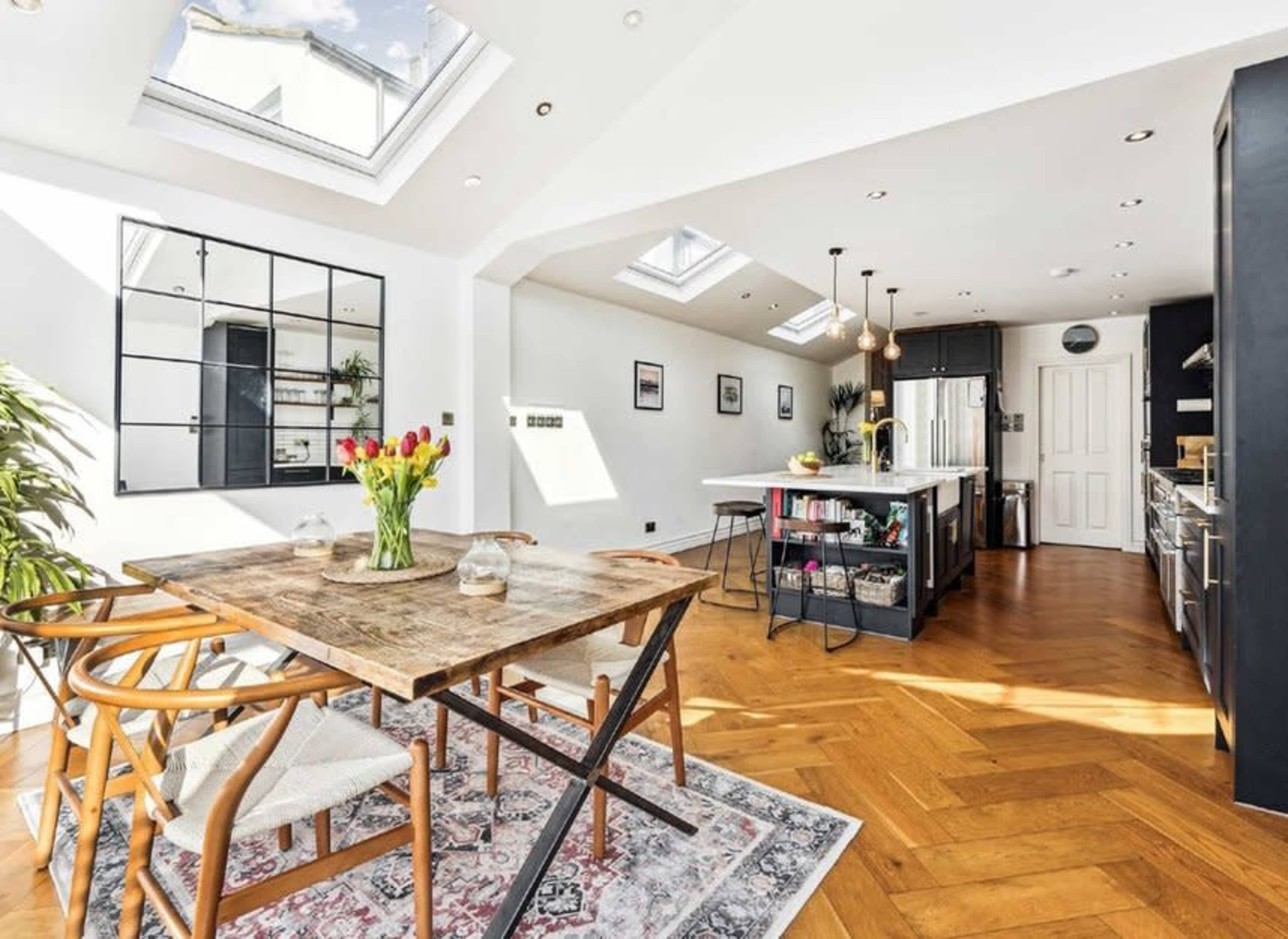 The image shows a modern kitchen and dining area featuring a wooden table with flowers, an island in the center, and skylights that provide ample natural light.