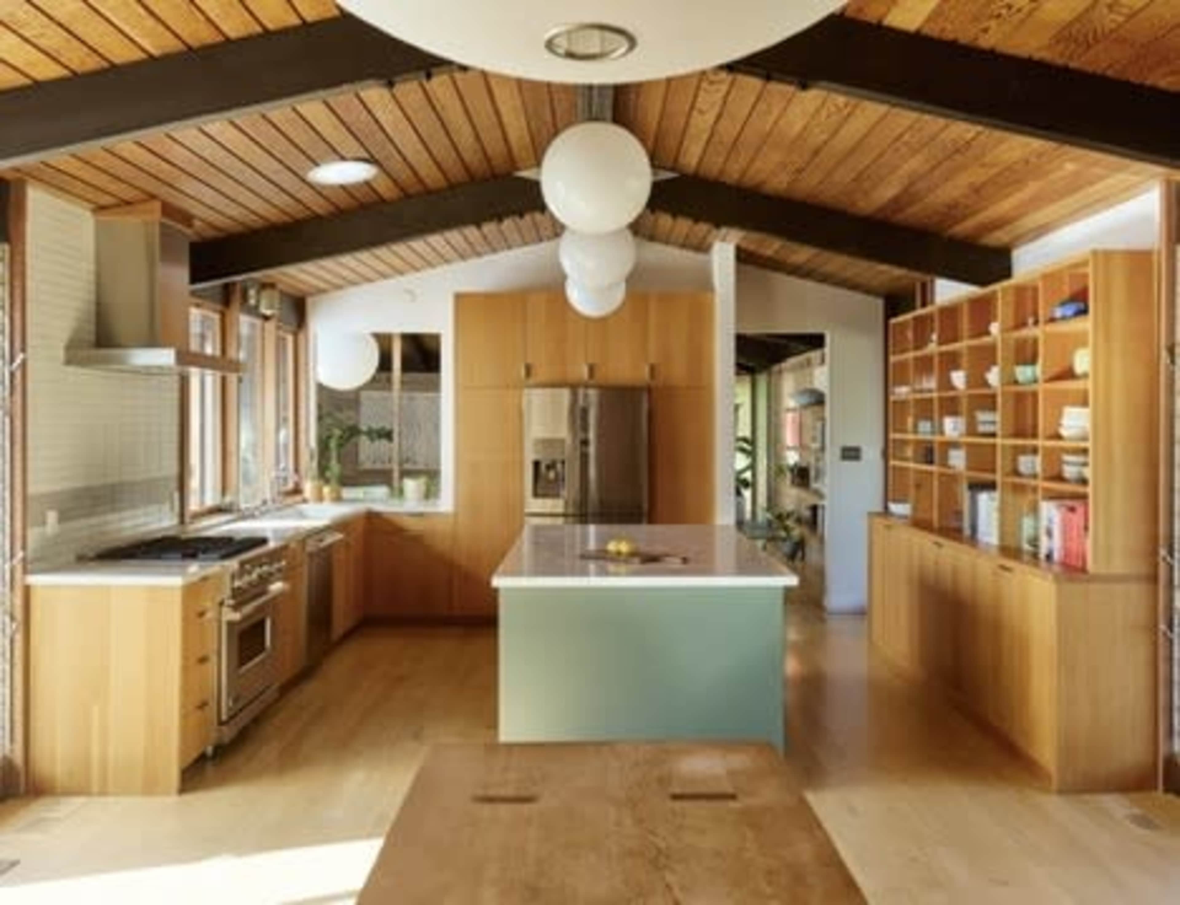 The image shows a modern kitchen featuring wooden cabinetry, a central island with a white countertop, and large windows allowing natural light to fill the space.