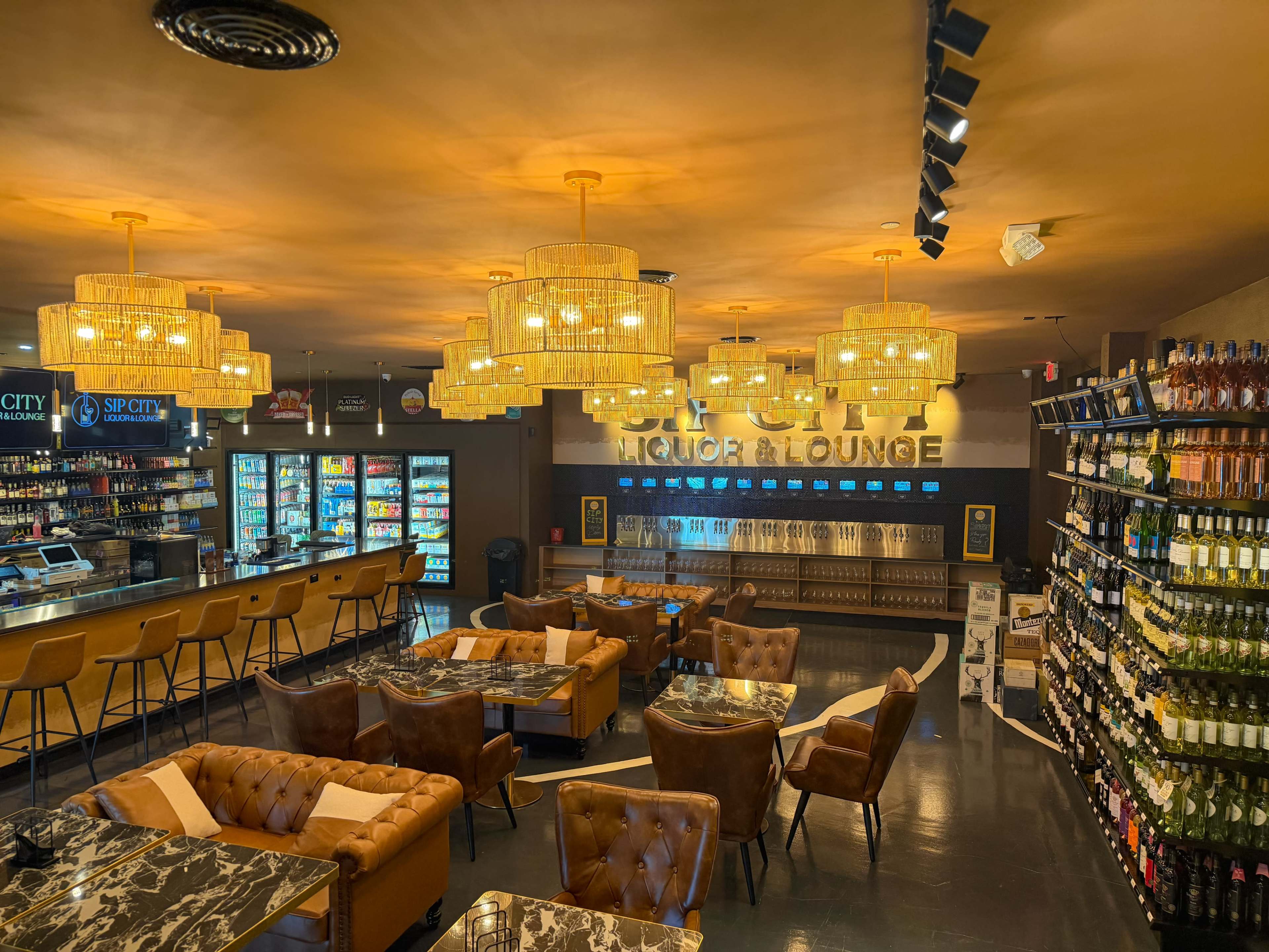 The image shows an interior of a liquor lounge featuring a bar area with shelves of beverages, comfortable seating, and multiple hanging light fixtures.