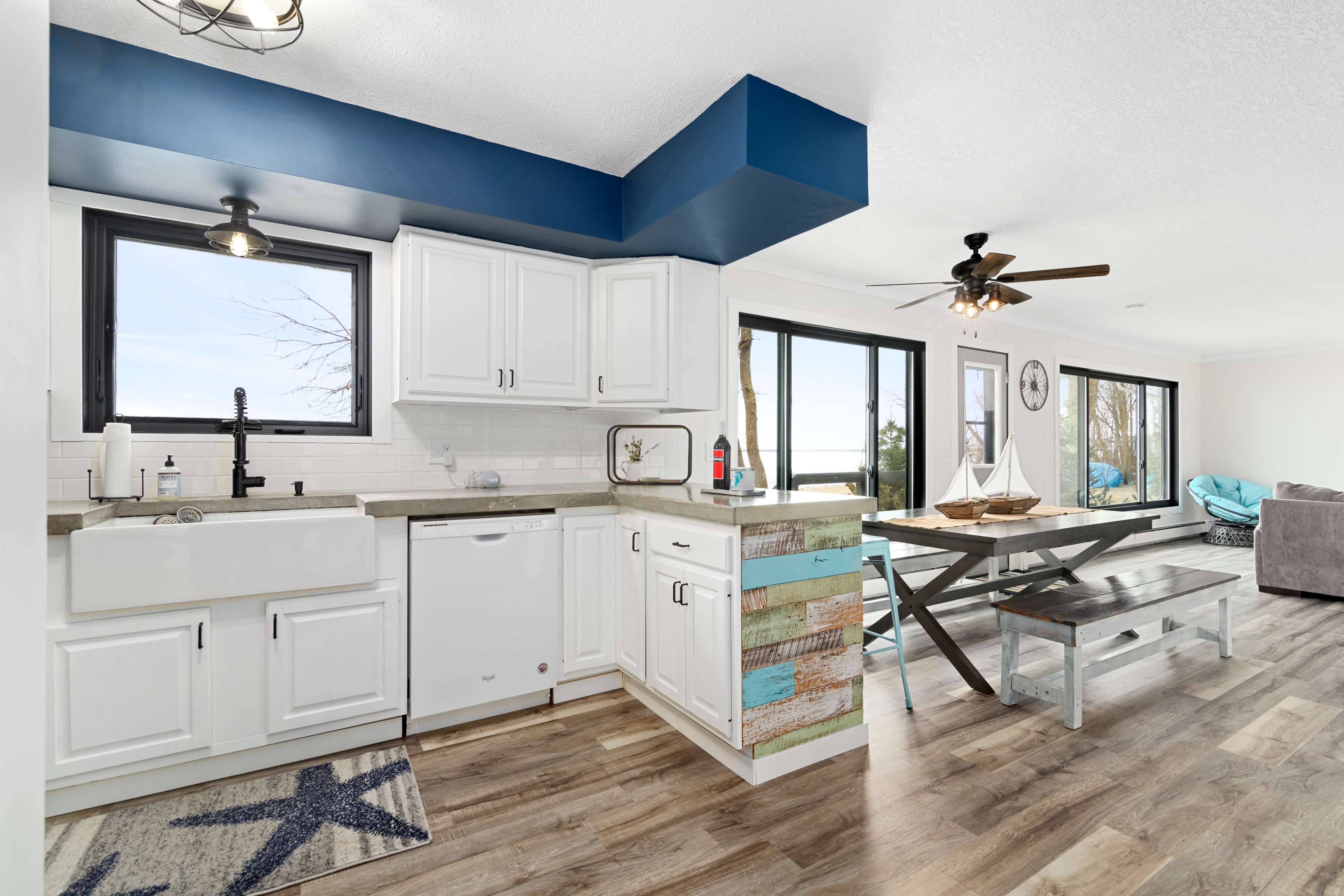 A bright, modern kitchen with white cabinets and a farmhouse sink, adjacent to a dining area featuring a picnic-style table and large windows overlooking a scenic view.
