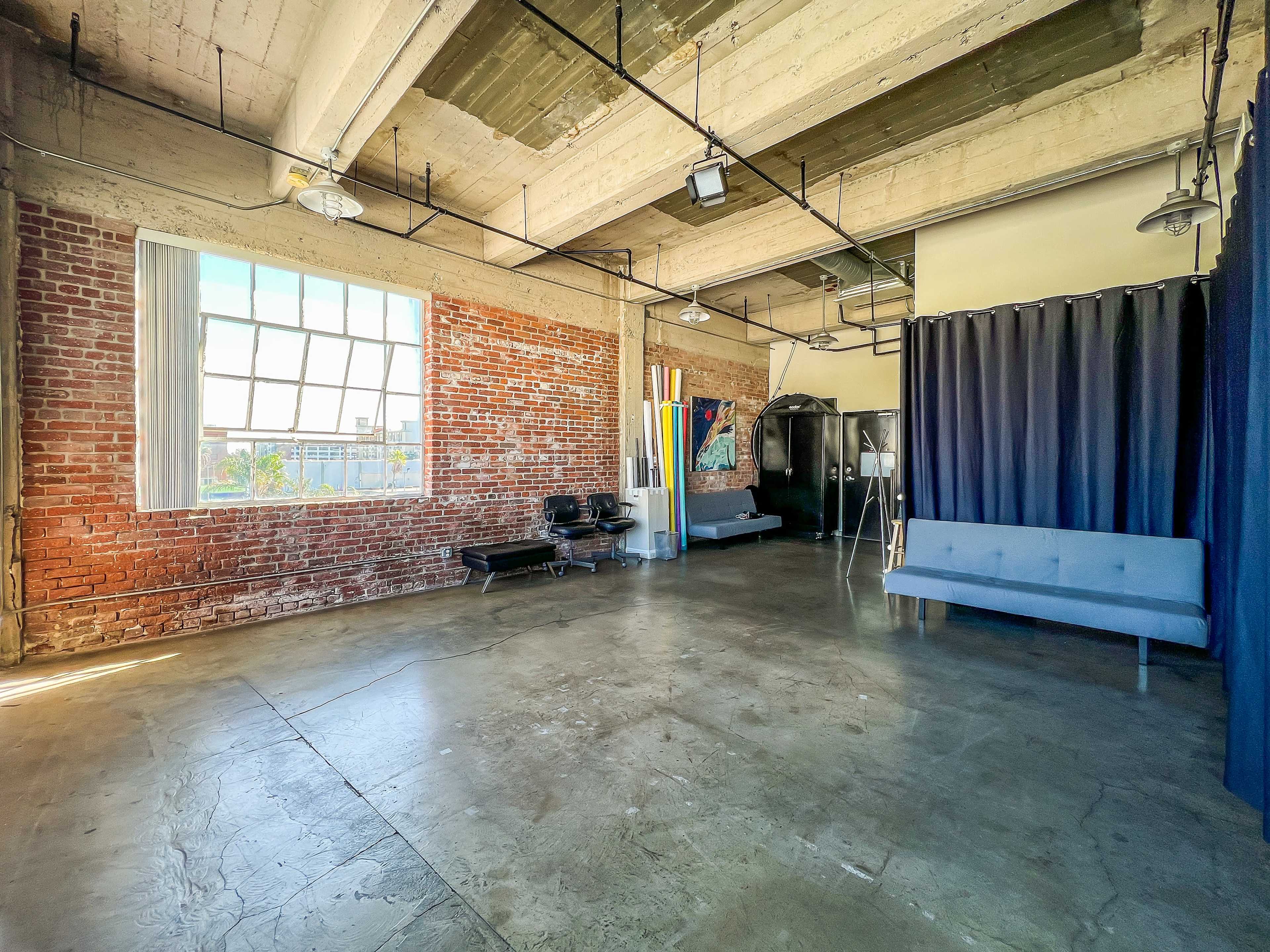 An industrial-style room with exposed brick walls, large windows, and minimal furniture including a gray sofa and black chairs.