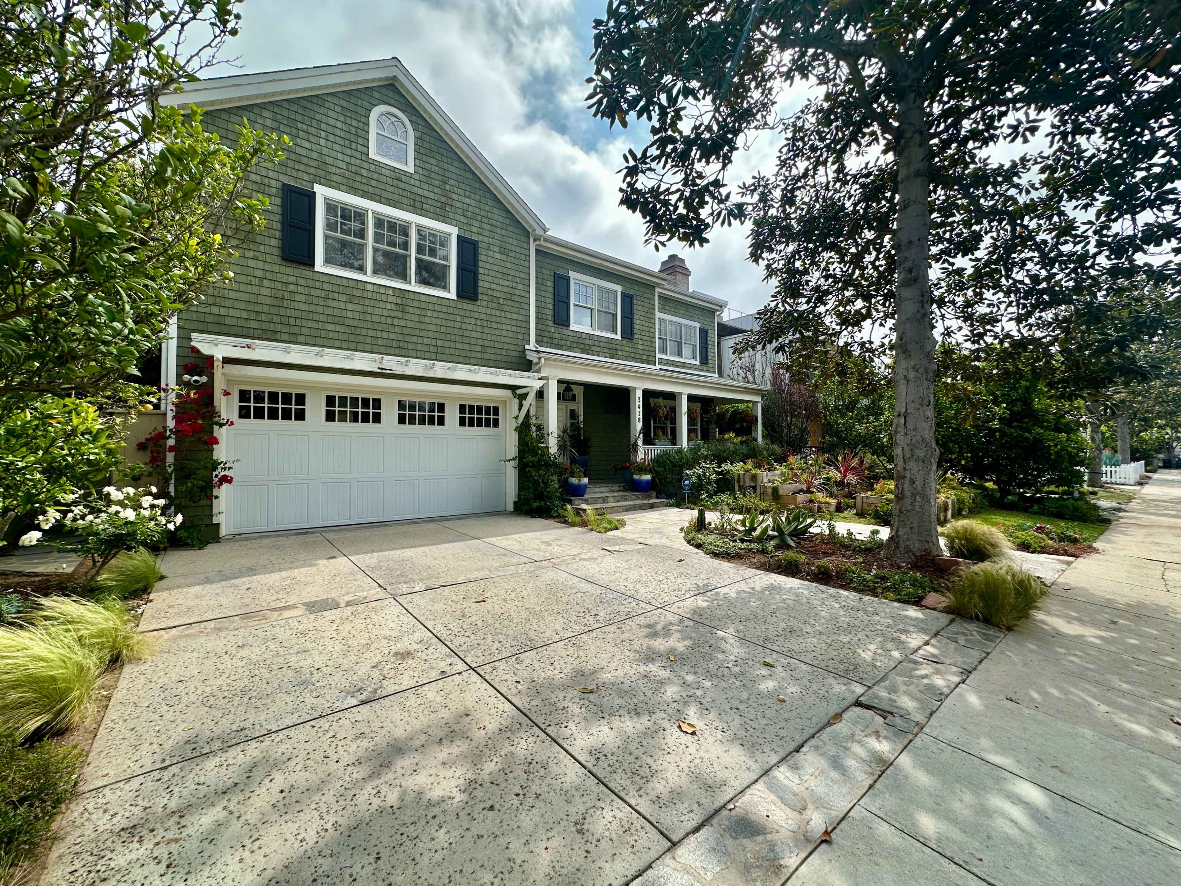 The image shows a large, green-shingled house with a garage and a well-maintained front yard featuring various plants and trees.